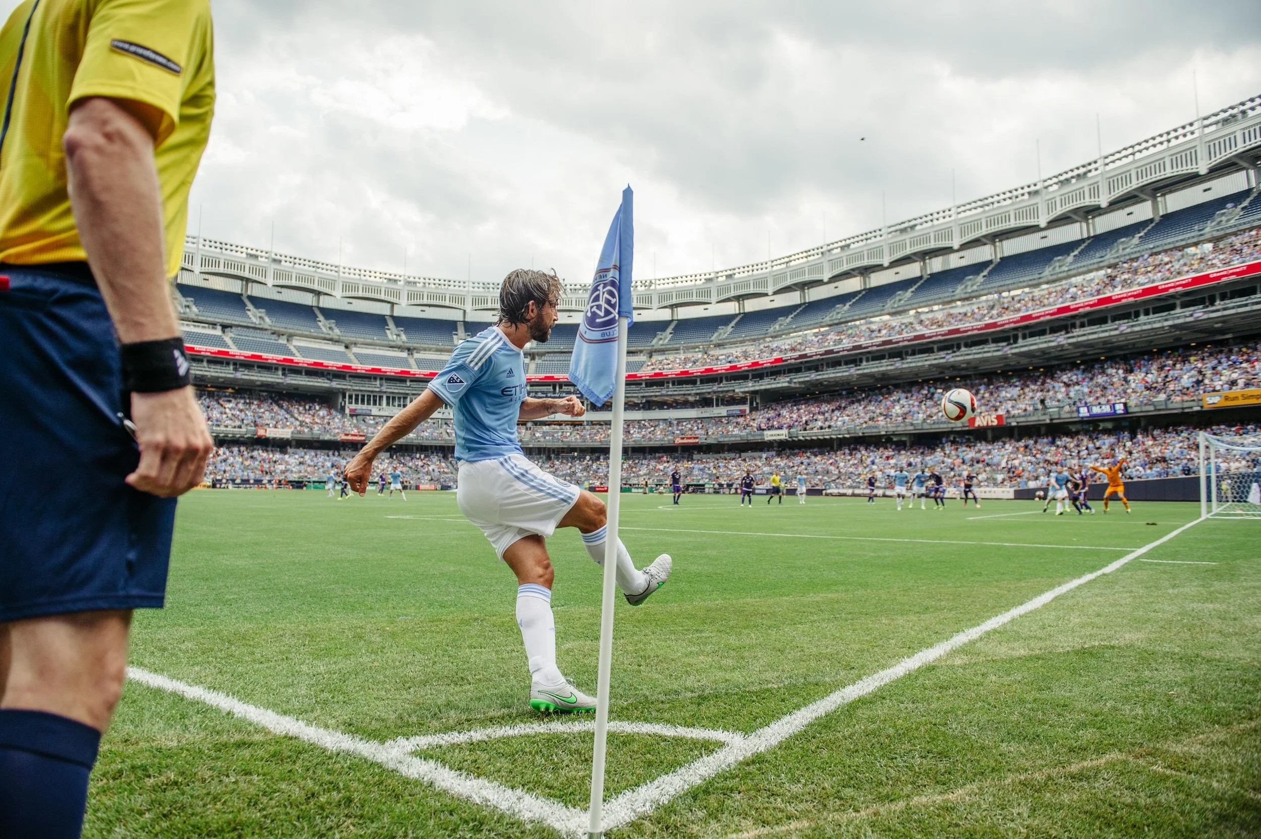A soccer player in a light blue uniform kicks the ball near the corner flag on a soccer field during a match, with a crowd in the stands and a referee visible on the left side of the image.