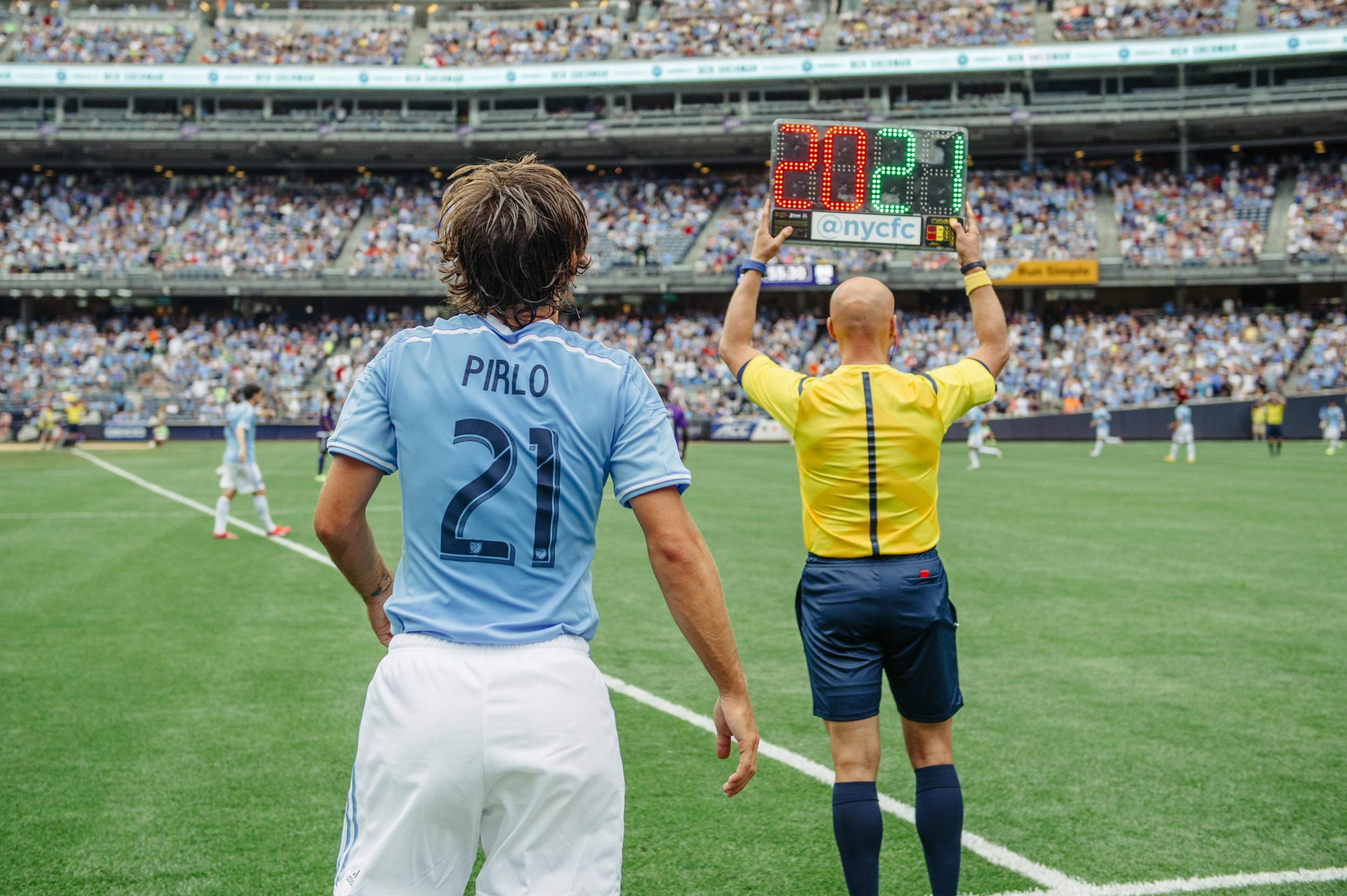 Soccer player wearing light blue jersey with the name 'Pirlo' and number 21 stands on the field while a referee in a yellow shirt holds a substitution board showing 20 and 21, with a crowd in the stands.