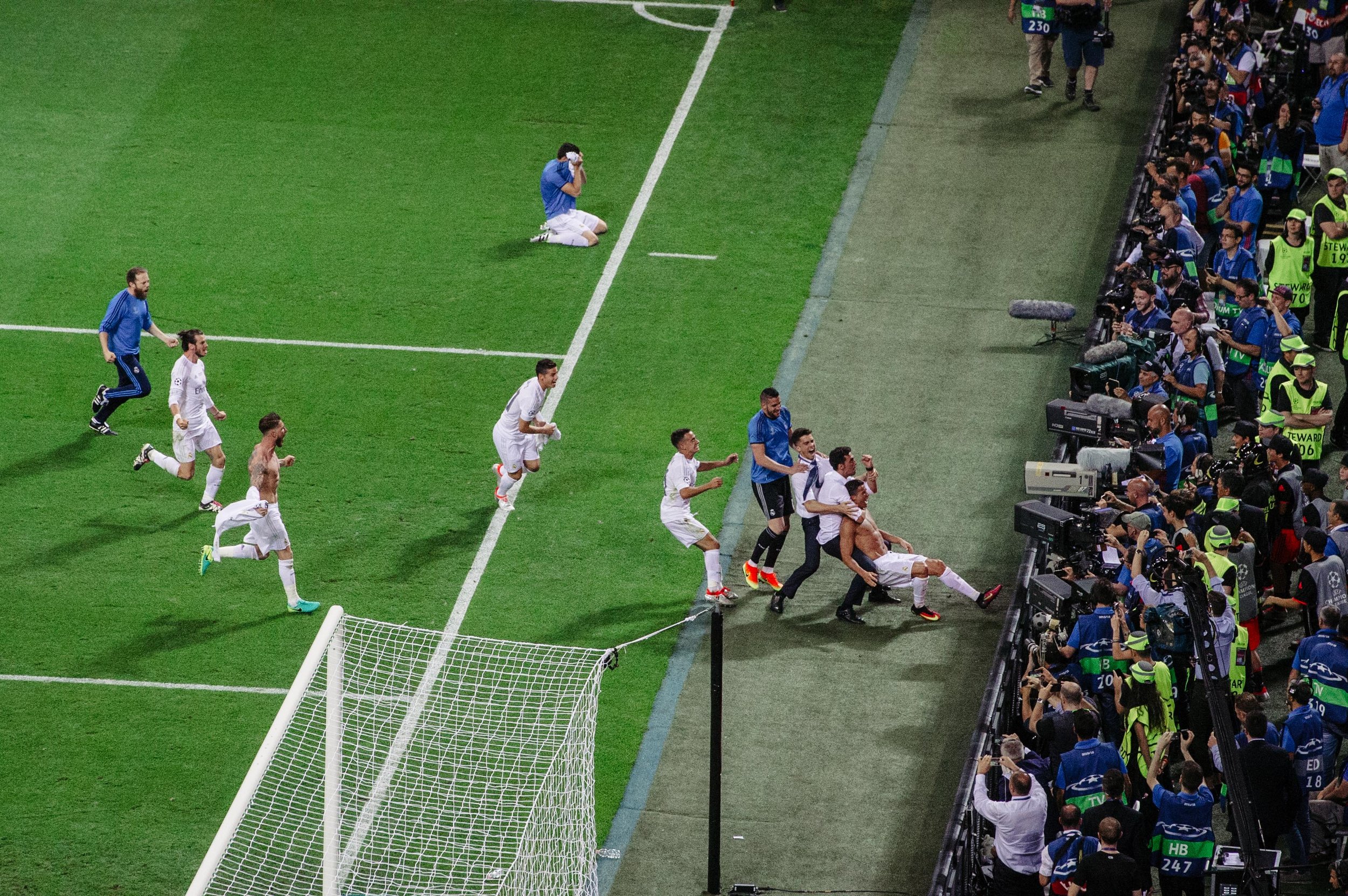 A soccer team celebrating a goal near the corner flag, with a large crowd and media personnel on the sidelines.
