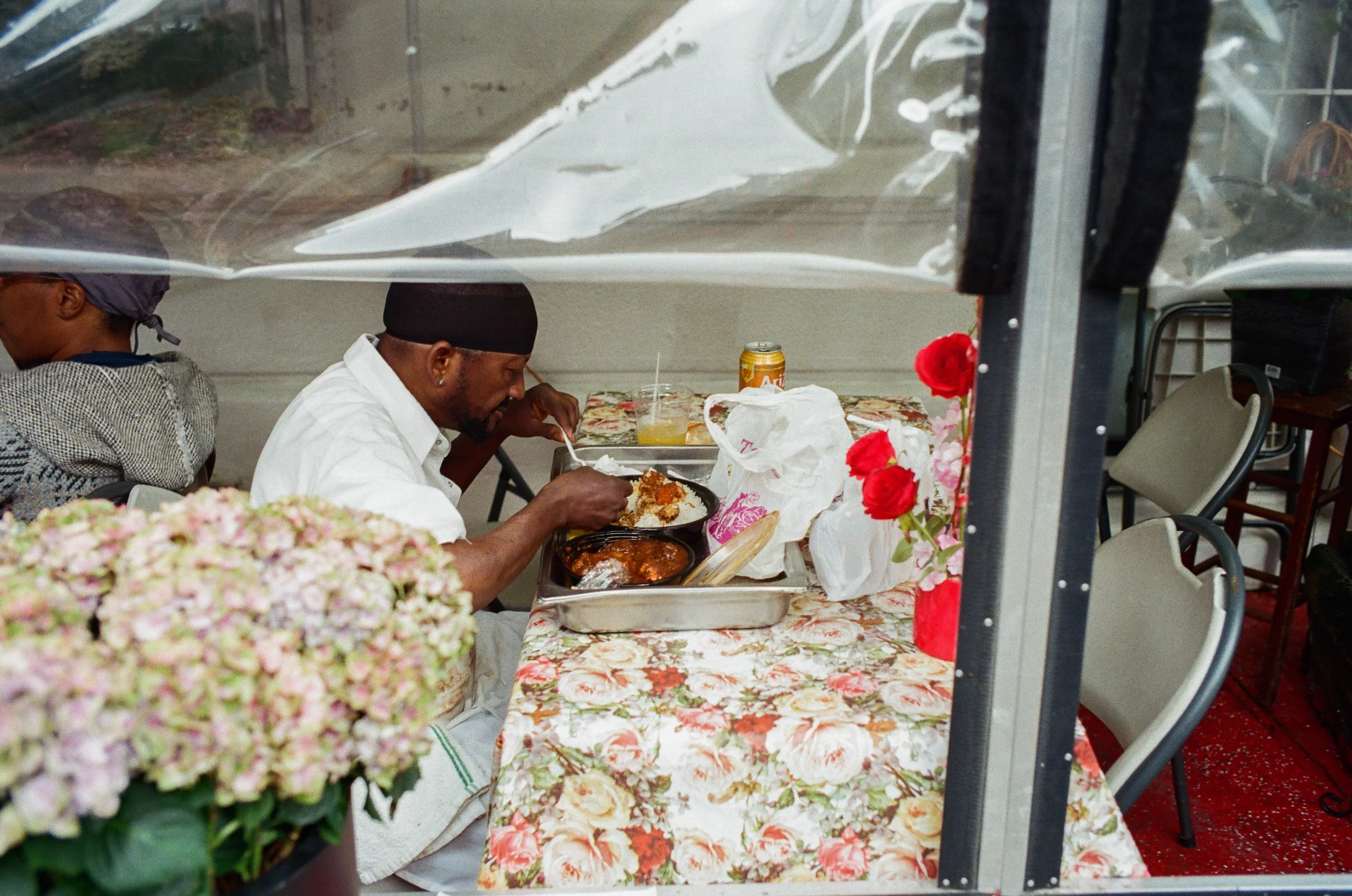 Man in white shirt and black head covering eating at a table with floral tablecloth, with flowers and chairs visible around.