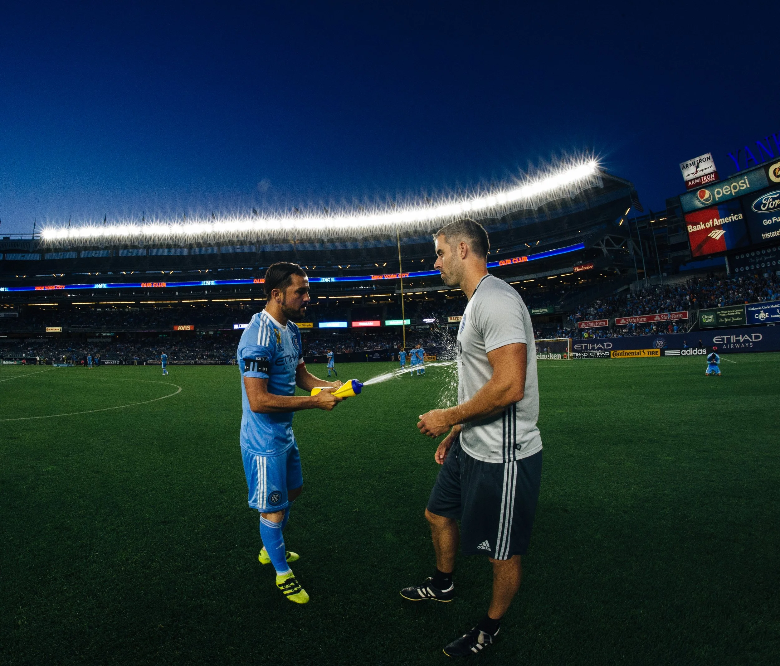 Two soccer players on a field, one in a blue uniform and the other in a gray shirt, with a crowd in the stadium background during dusk or evening.