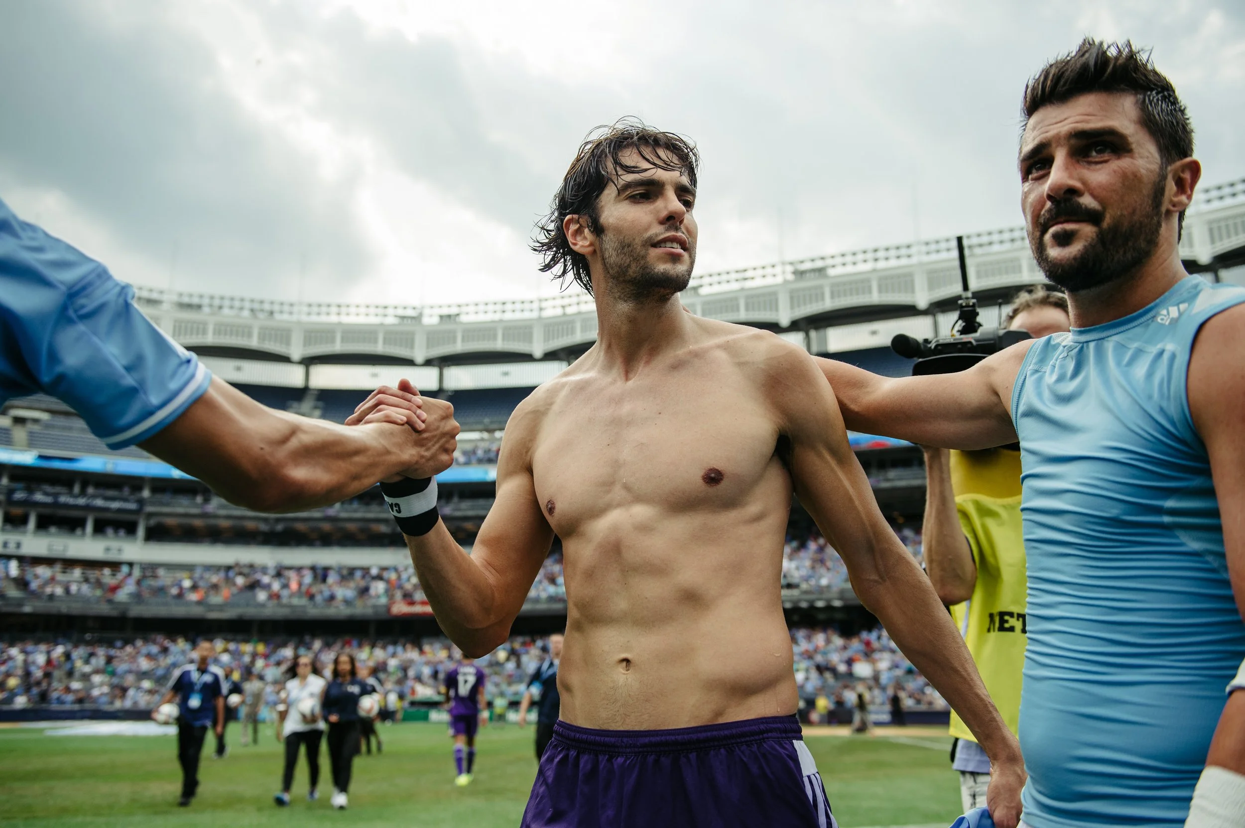 Two soccer players, one shirtless, shake hands on the field. The shirtless player is between two men in blue shirts.