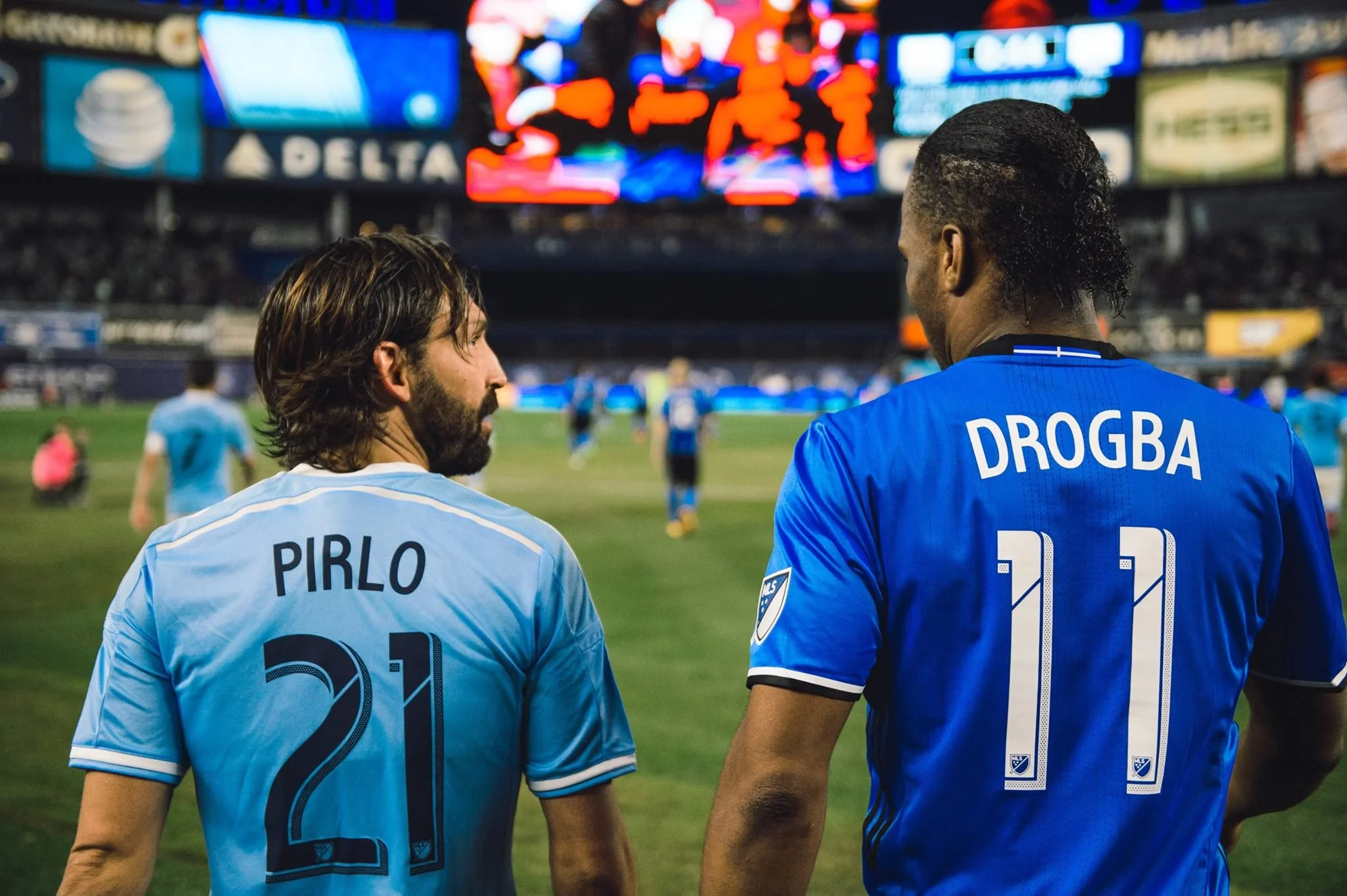 Two soccer players face away from the camera on a field, one wearing a light blue jersey with 'Pirlo' and the number 21, and the other wearing a dark blue jersey with 'Drogba' and the number 11, with a stadium and scoreboard in the background.