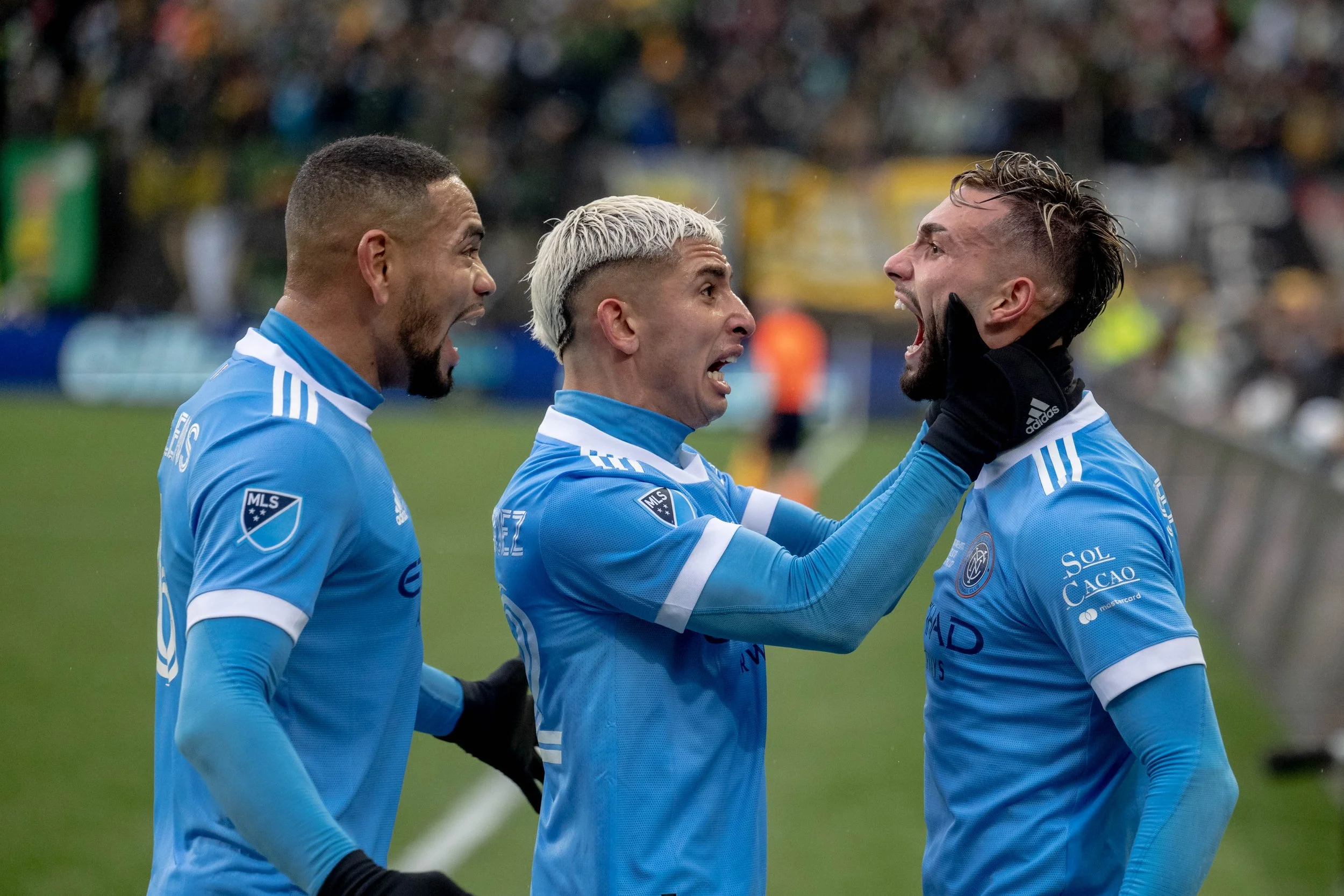 Three soccer players in blue jerseys celebrating on the field, with two of them shouting at each other and the middle player holding the face of the player on the right.