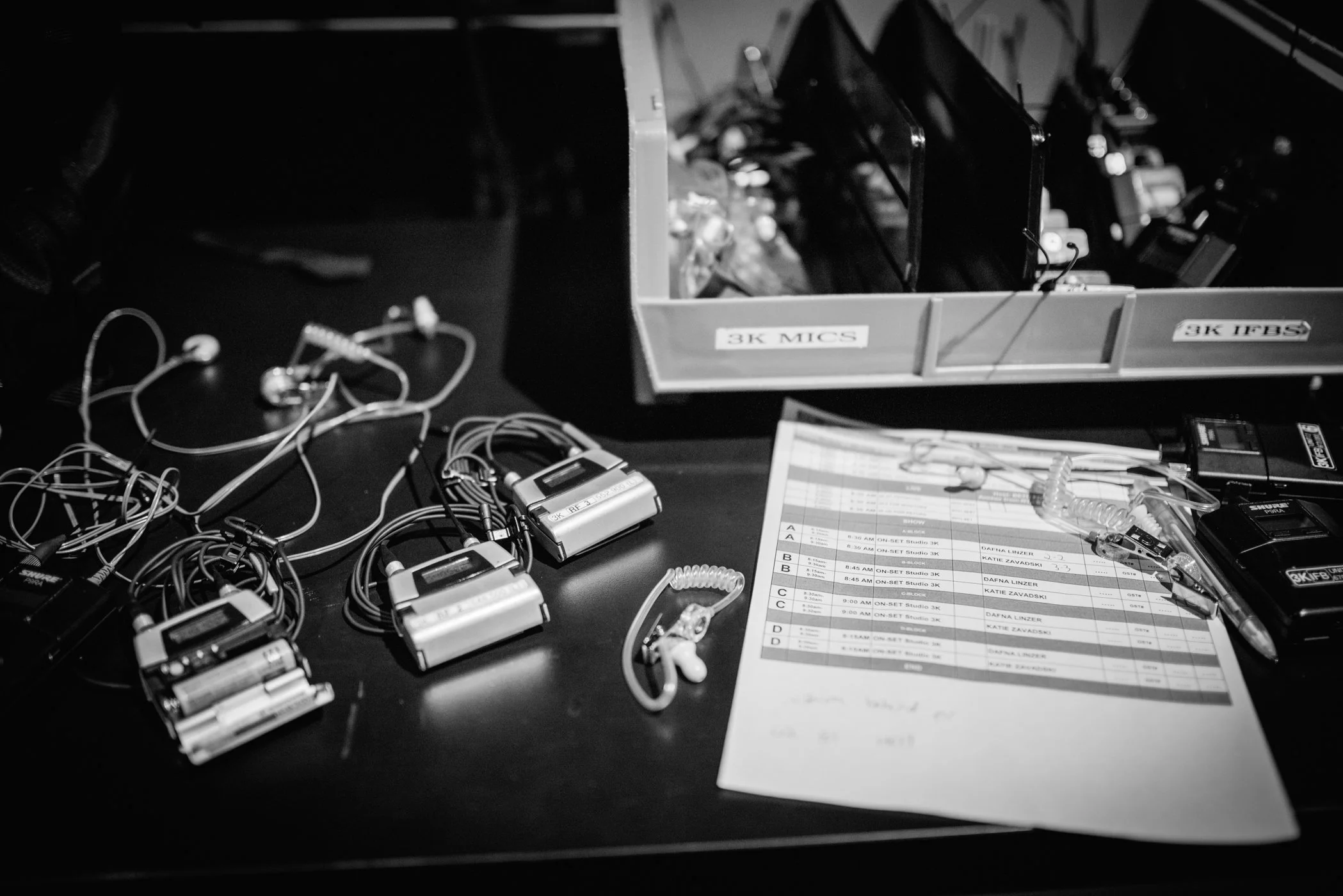 A collection of wireless microphone transmitters, earbuds, and a clipboard with a schedule on a black table, with a plastic bin labeled '3K MICS' containing microphone receivers and accessories.