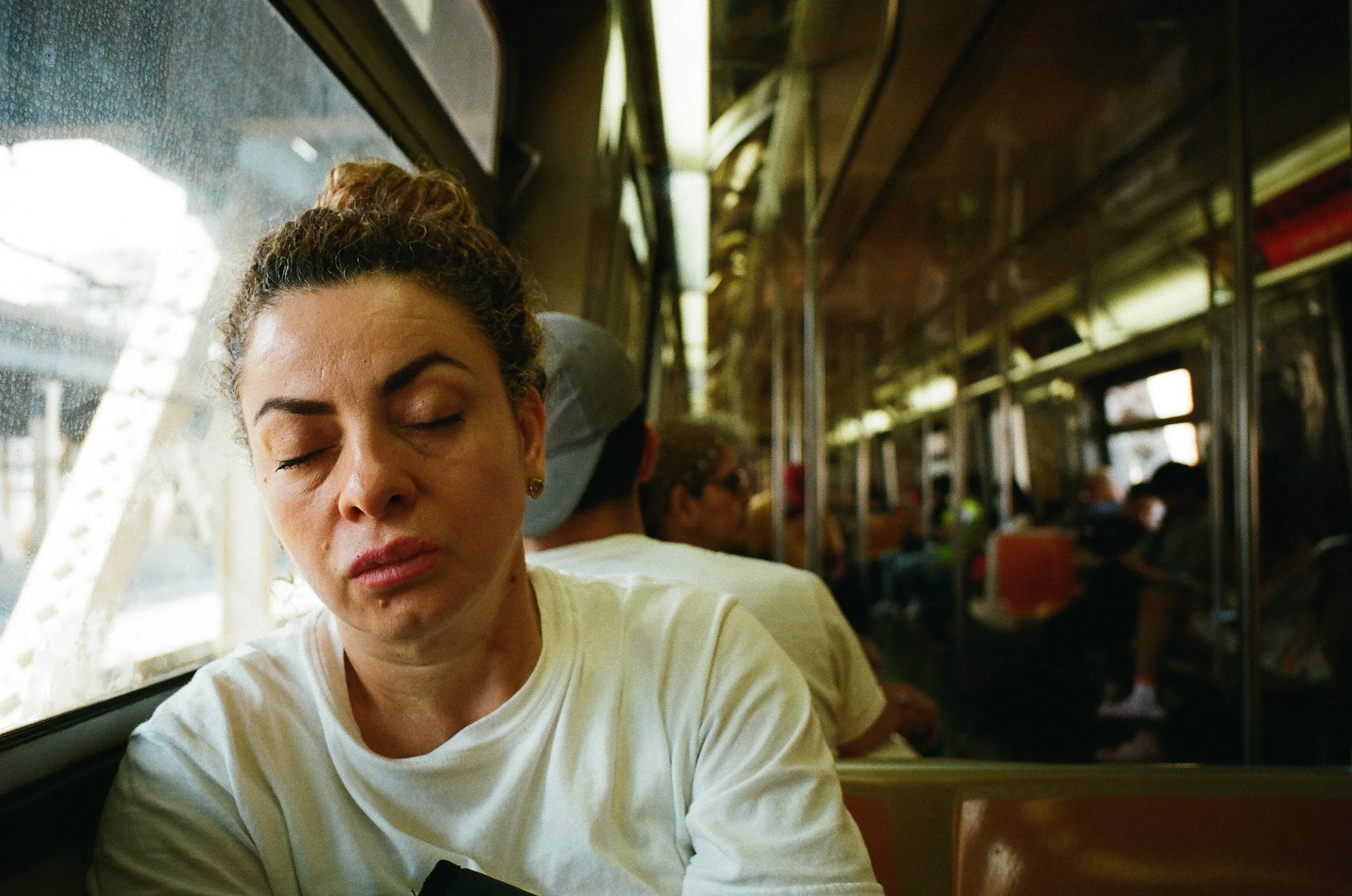 A woman with closed eyes sitting on a bus or train, with other passengers seated behind her, and the window showing a blurry exterior.