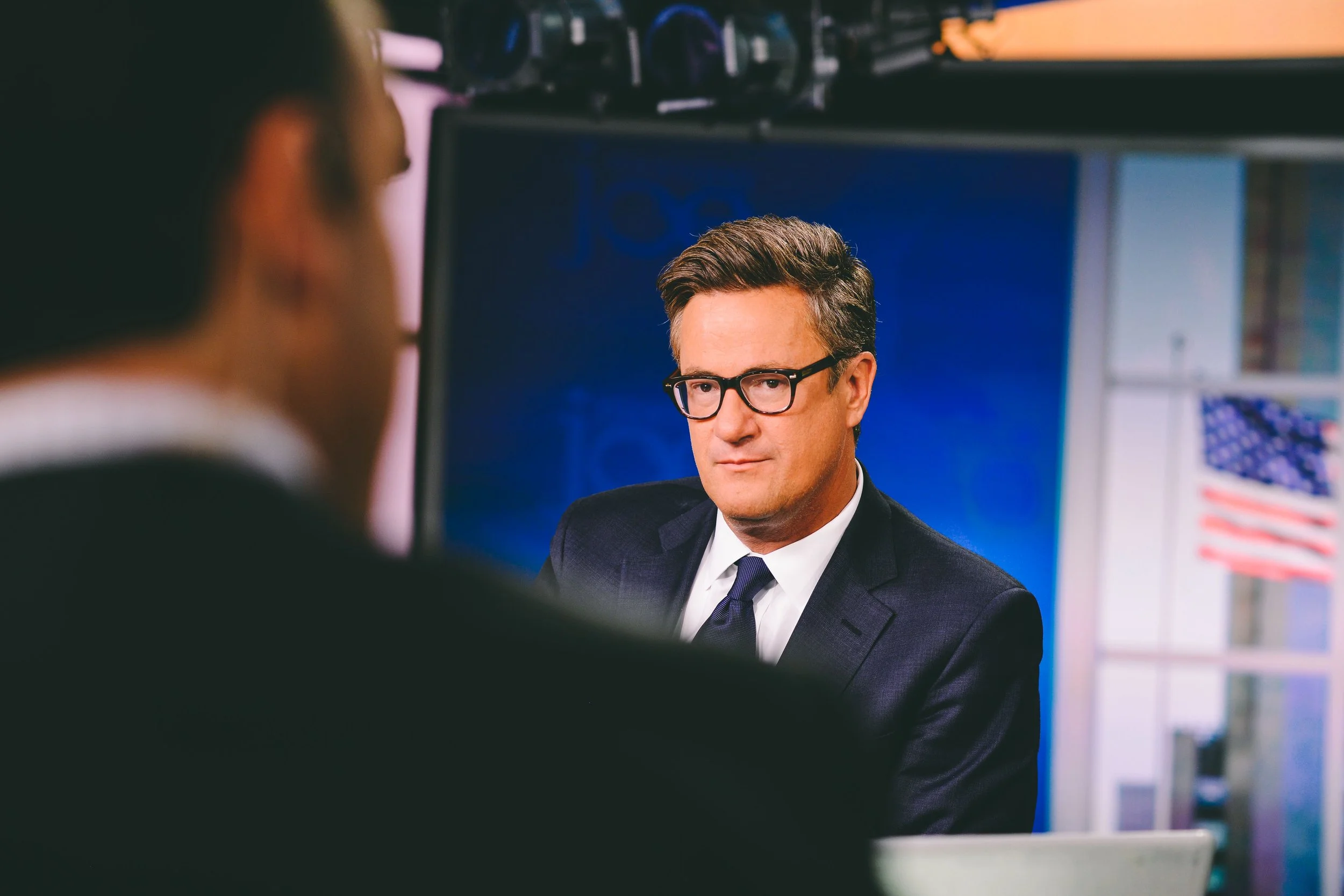 Man in glasses and suit sitting at a desk in front of a blue backdrop with an American flag in the background.
