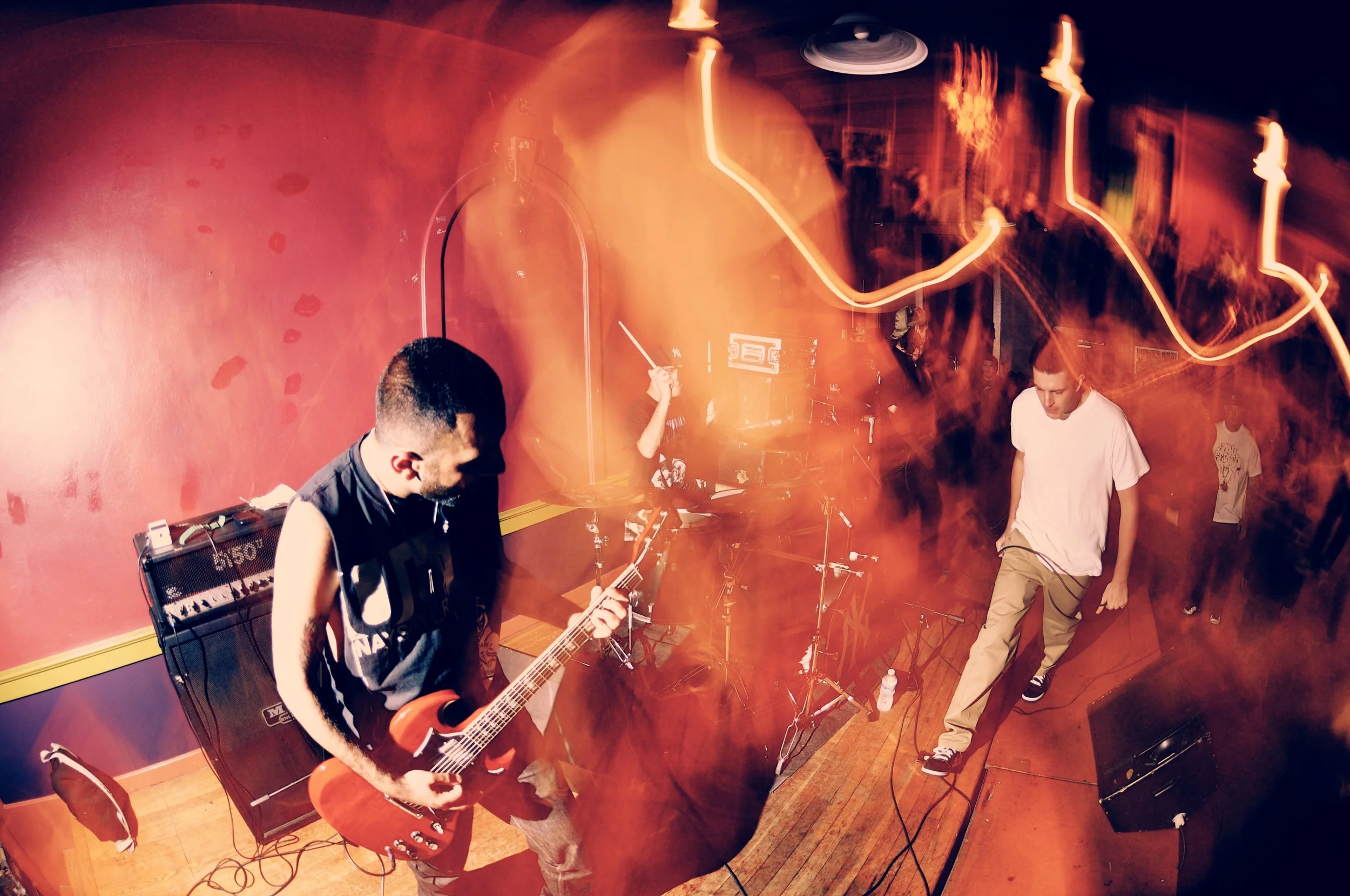 A band performing on stage with a guitarist, a drummer, and a vocalist in front of a red wall, with orange and yellow light trails creating a dynamic effect.