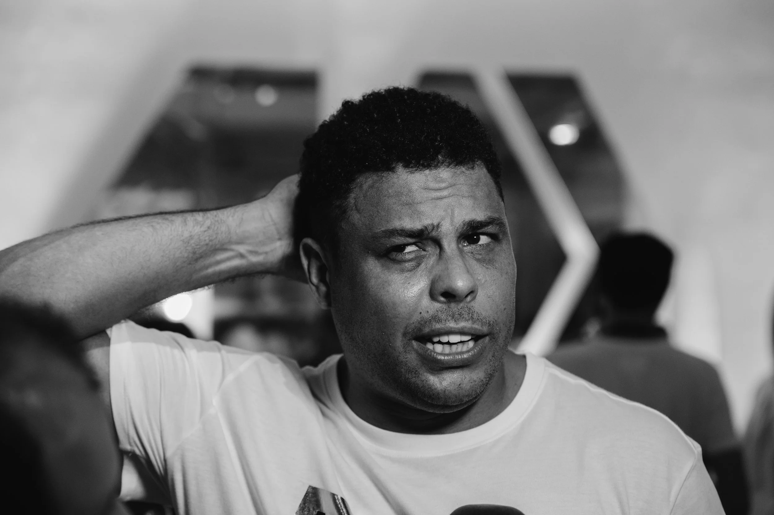 A man with short curly hair touching the back of his head, making a confused or annoyed facial expression in a black-and-white photo.