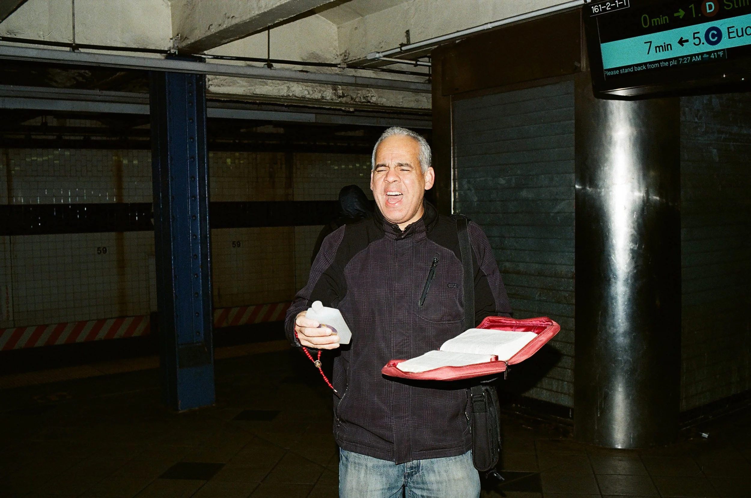 A man with gray hair, wearing a dark jacket, holding a red folder and a water bottle, standing in a subway station with a sign displaying subway directions and times.
