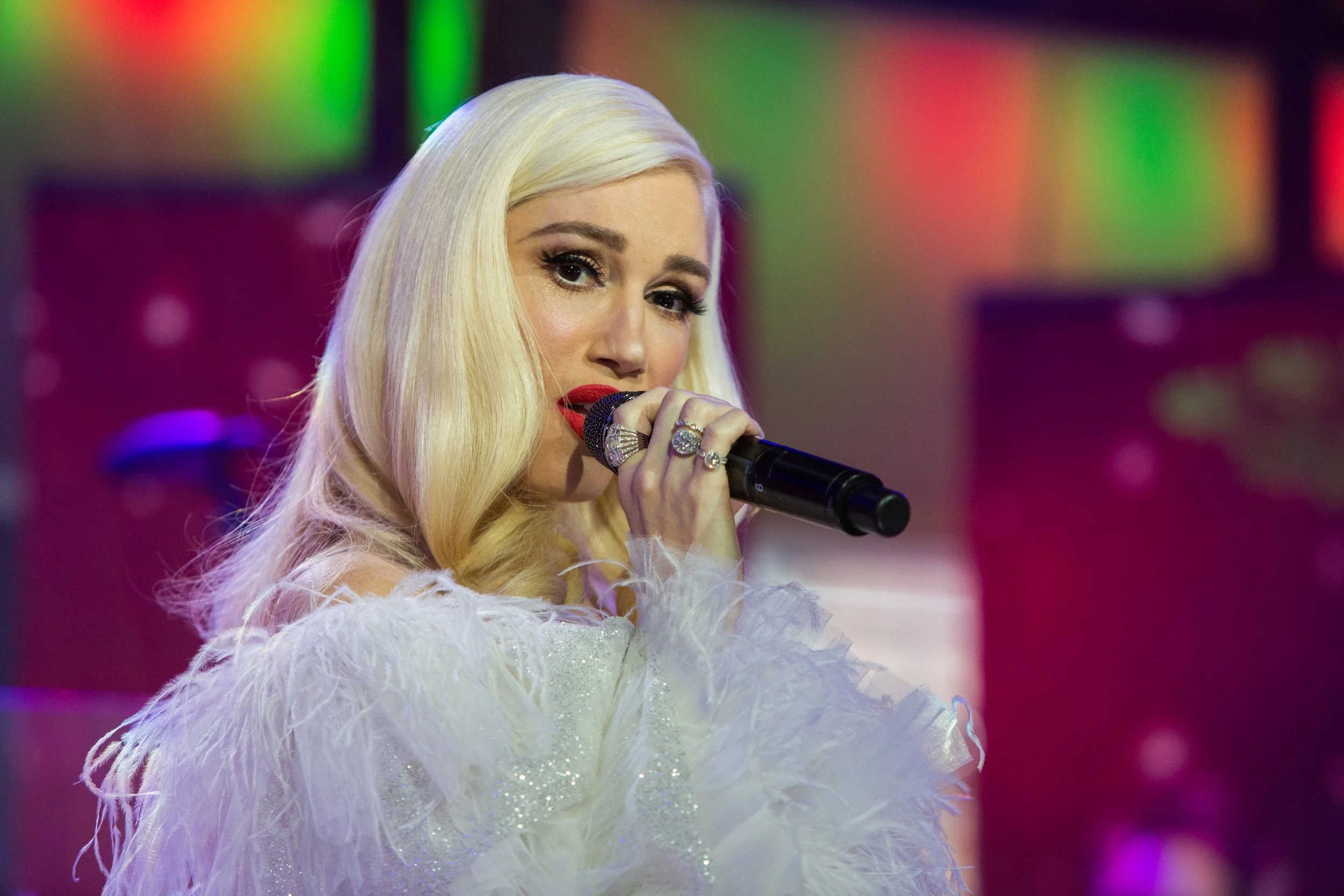 A blonde woman with red lipstick, wearing a white, feathered outfit, holds a microphone and performs on stage with colorful, blurred background lights.