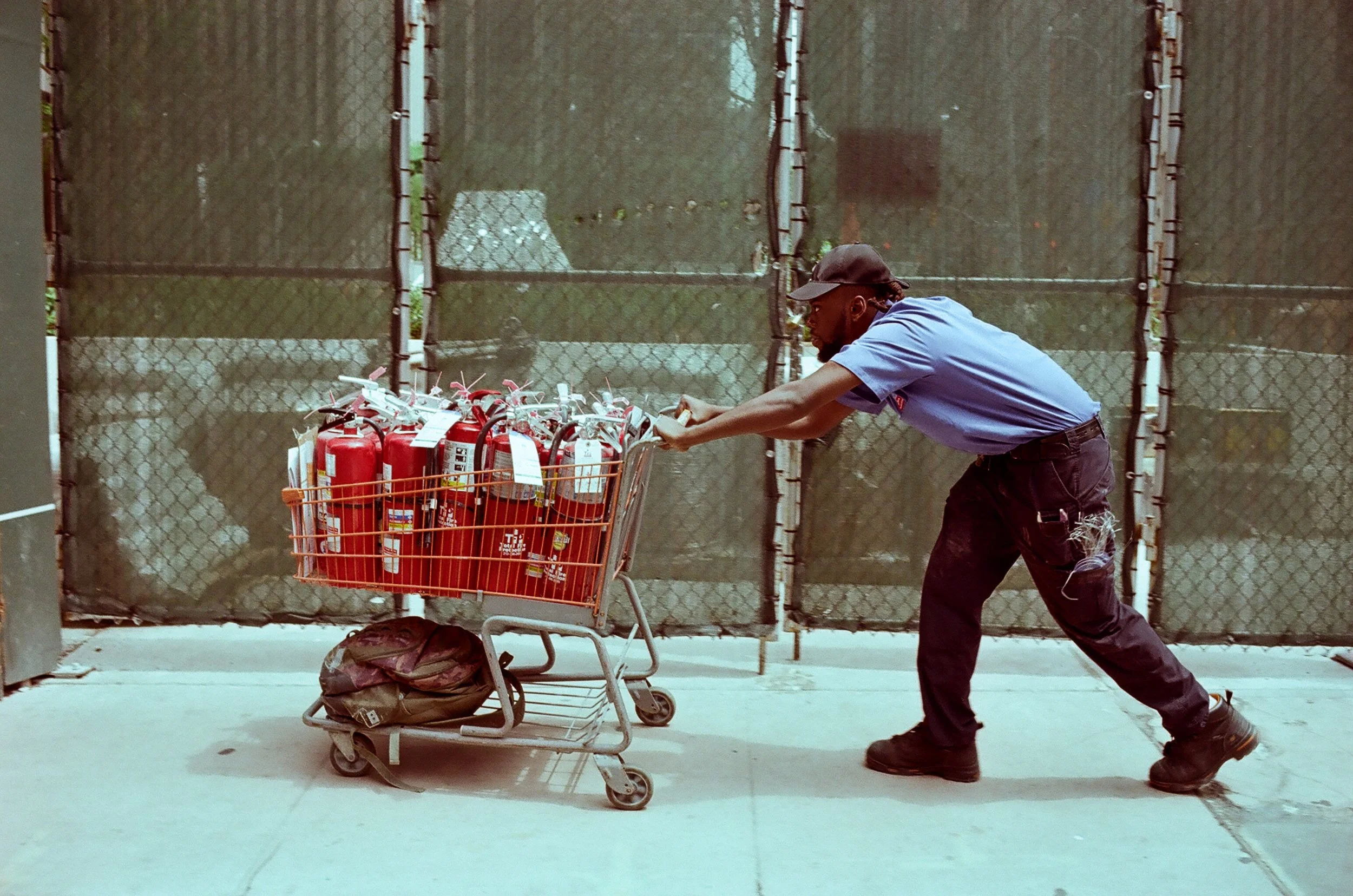 A man in a blue uniform and black cap pushing a shopping cart filled with multiple fire extinguishers on a sidewalk in front of a chain-link fence.