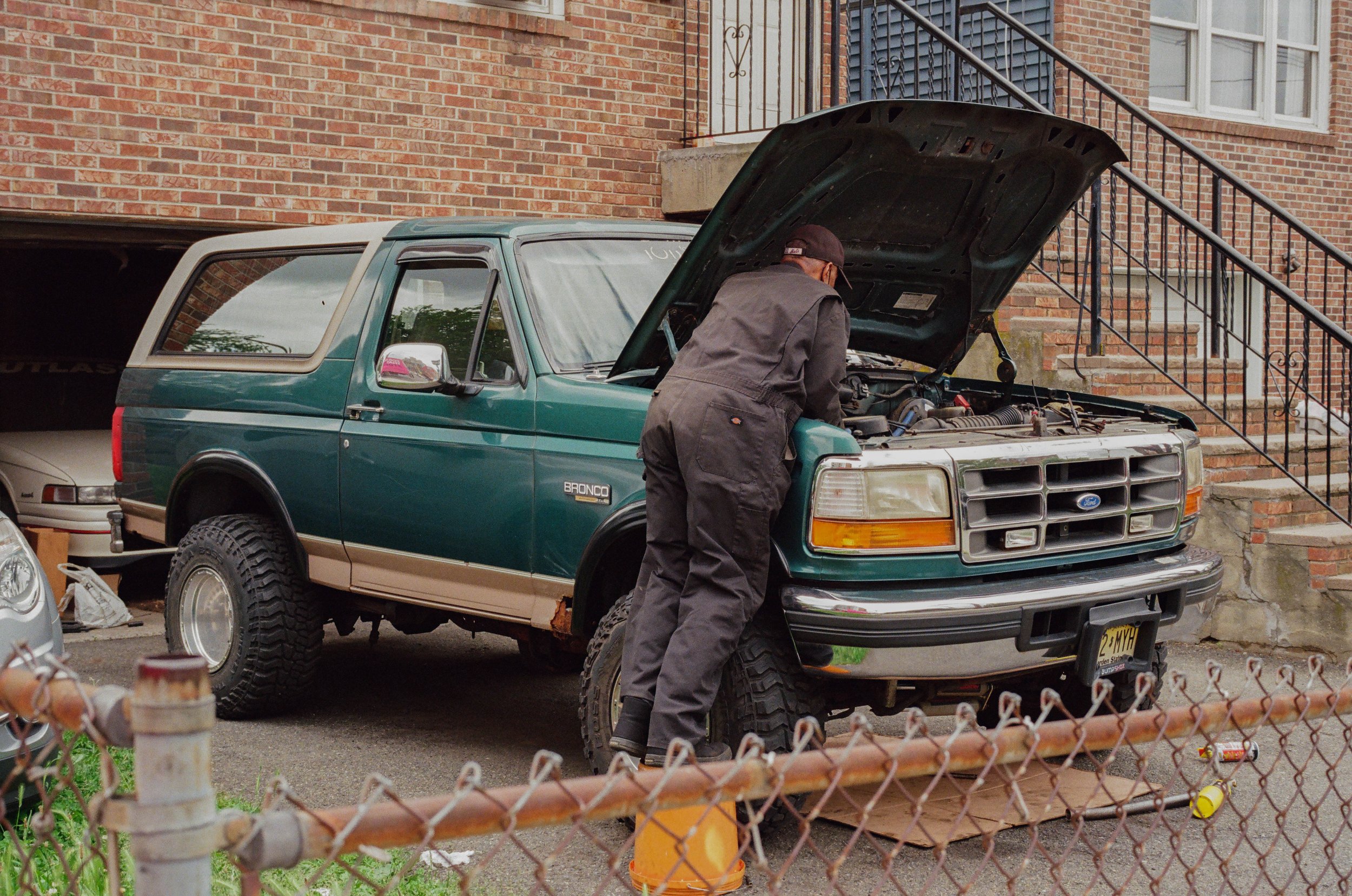 A person inspecting the engine of a teal Ford Bronco parked outside a brick building, with its hood open.