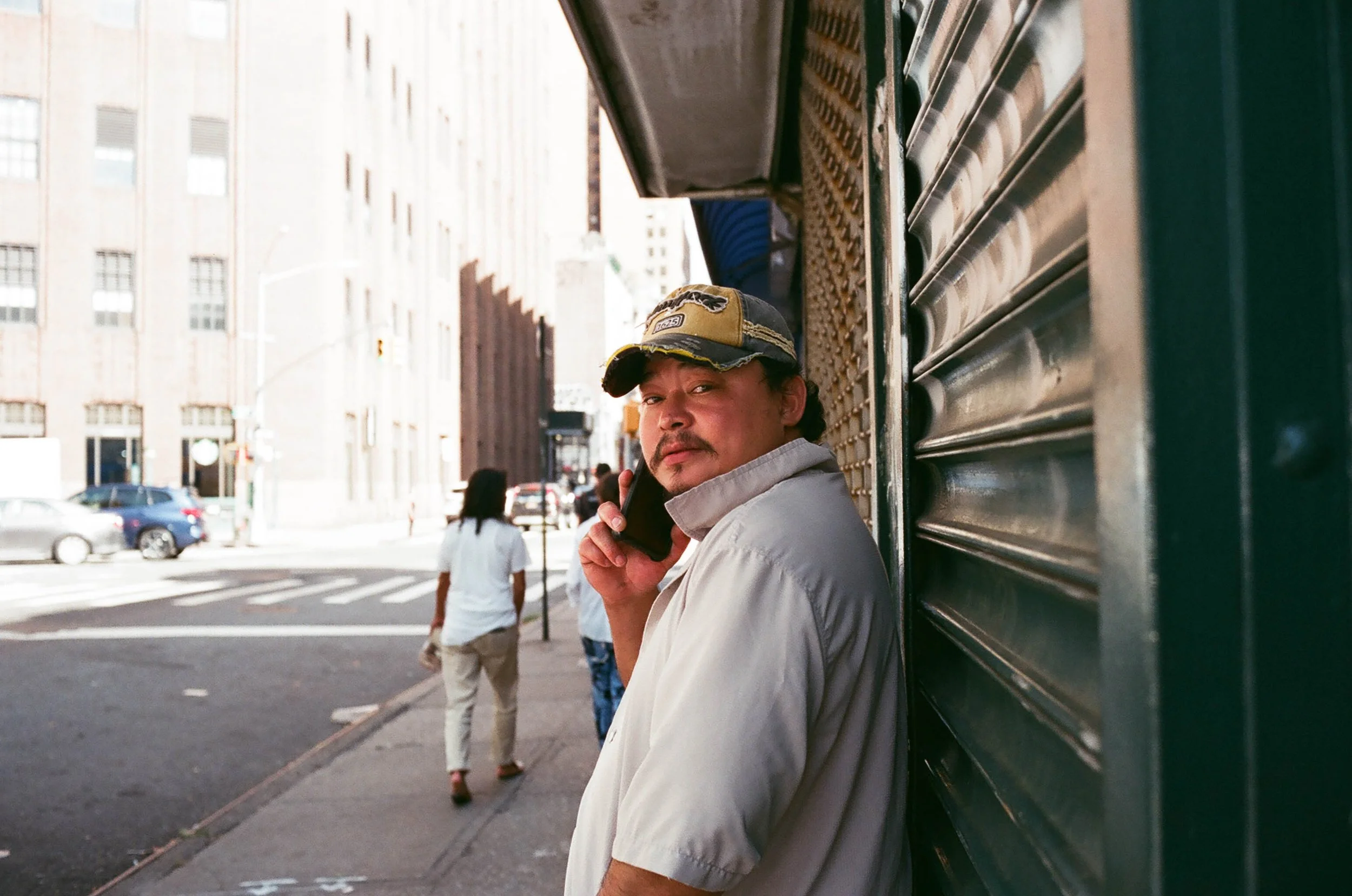 Man leaning against a green storefront talking on a cellphone on a city sidewalk, with pedestrians and cars in the background.