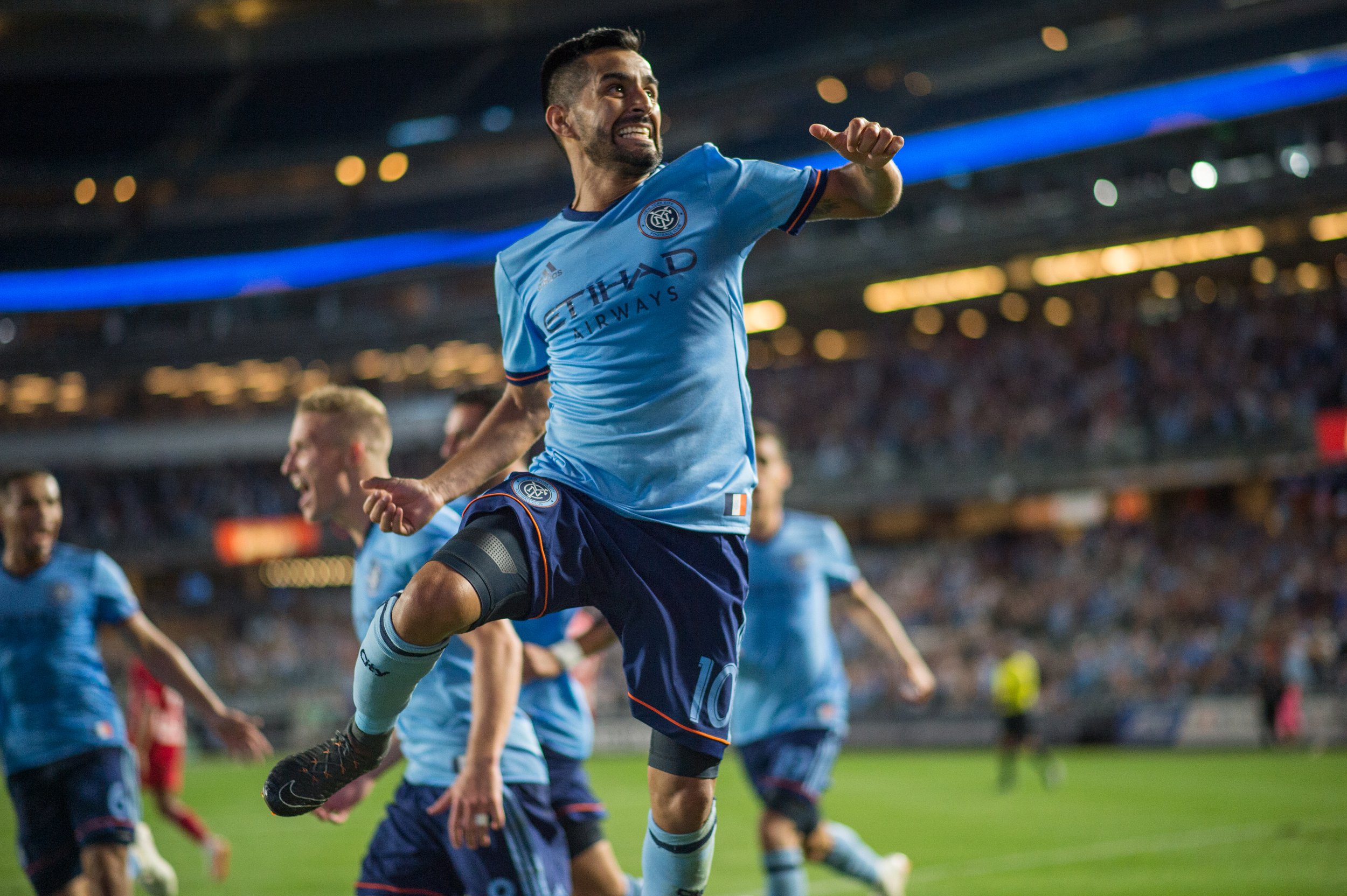 A soccer player celebrating a goal on the field with teammates, wearing a light blue jersey, at a stadium filled with spectators.