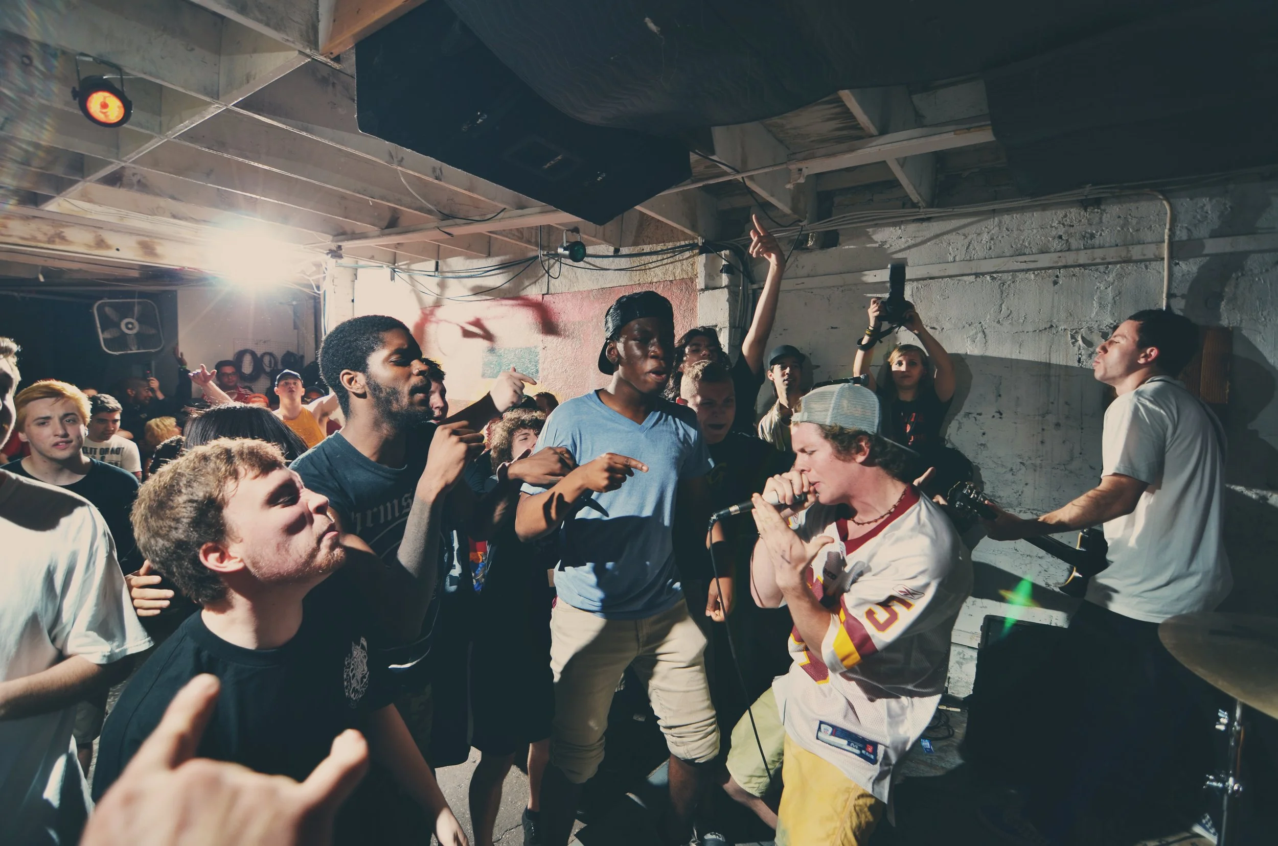 A live music performance in a small venue with a vocalist singing into a microphone, surrounded by a crowd of concertgoers dancing and raising their hands.