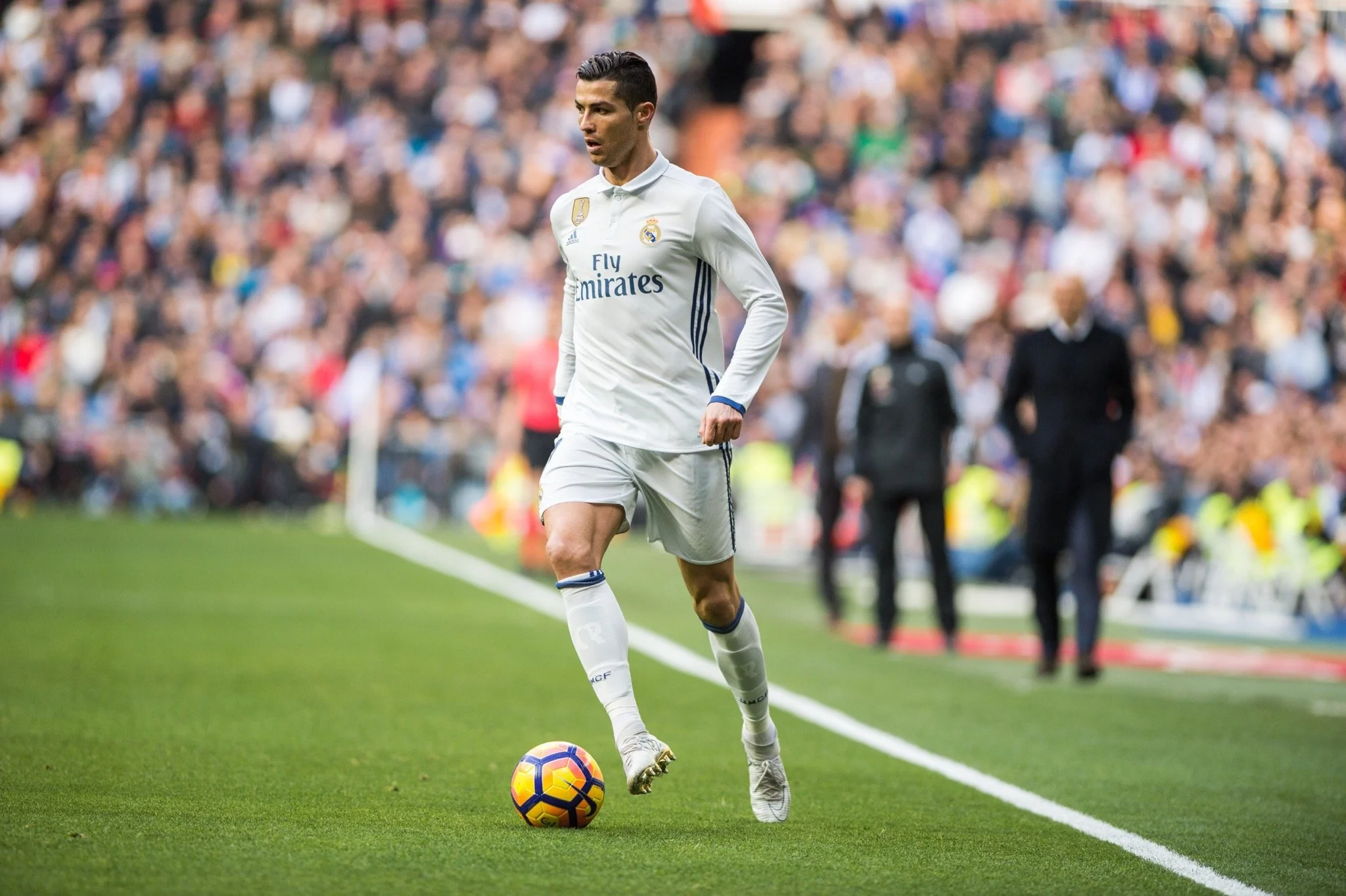 A soccer player, Cristiano Ronaldo, standing on the field in a white uniform with a soccer ball at his feet during a match with a large crowd in the background.