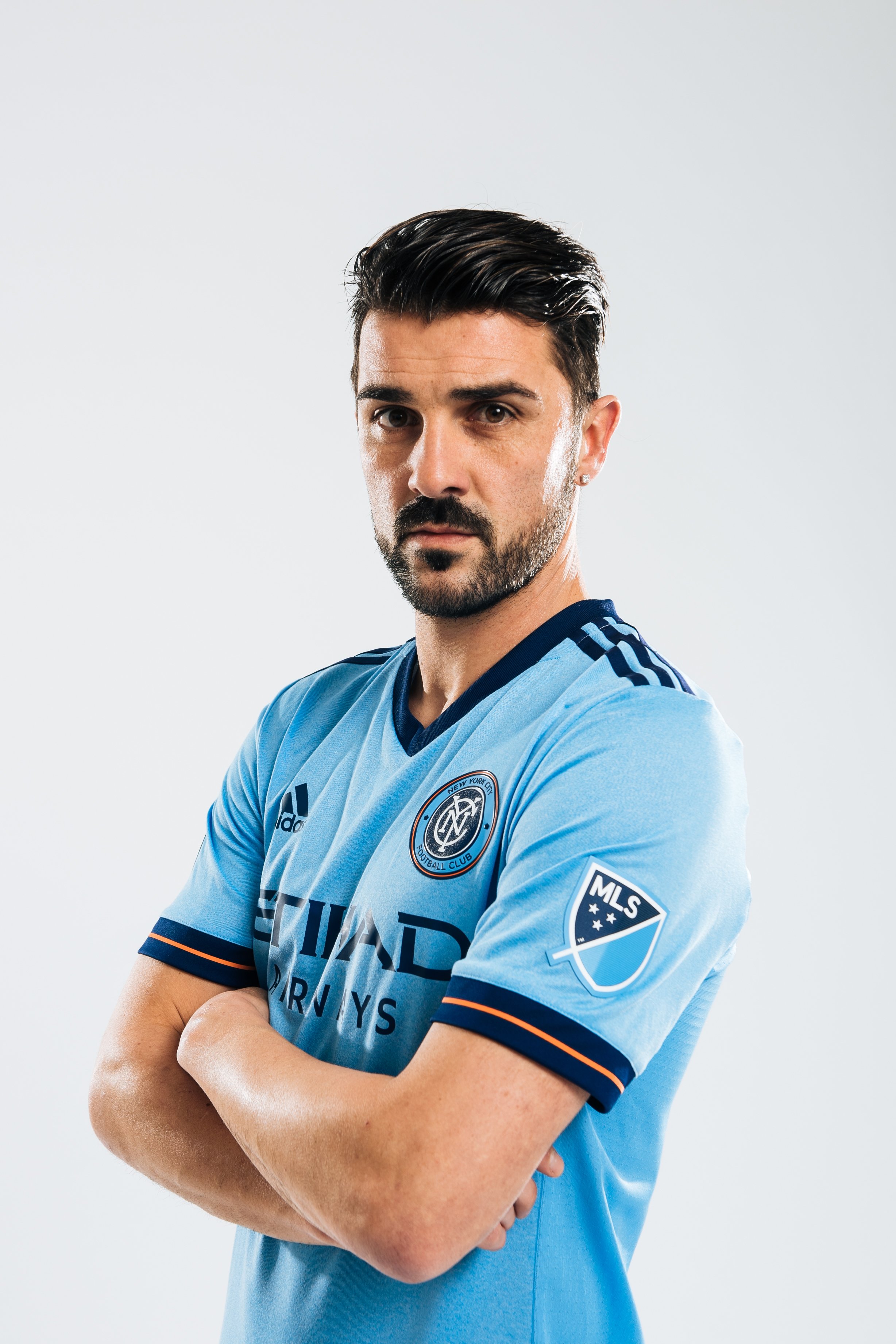 Portrait of a male soccer player in a light blue New York City Football Club jersey, standing with arms crossed against a plain white background.