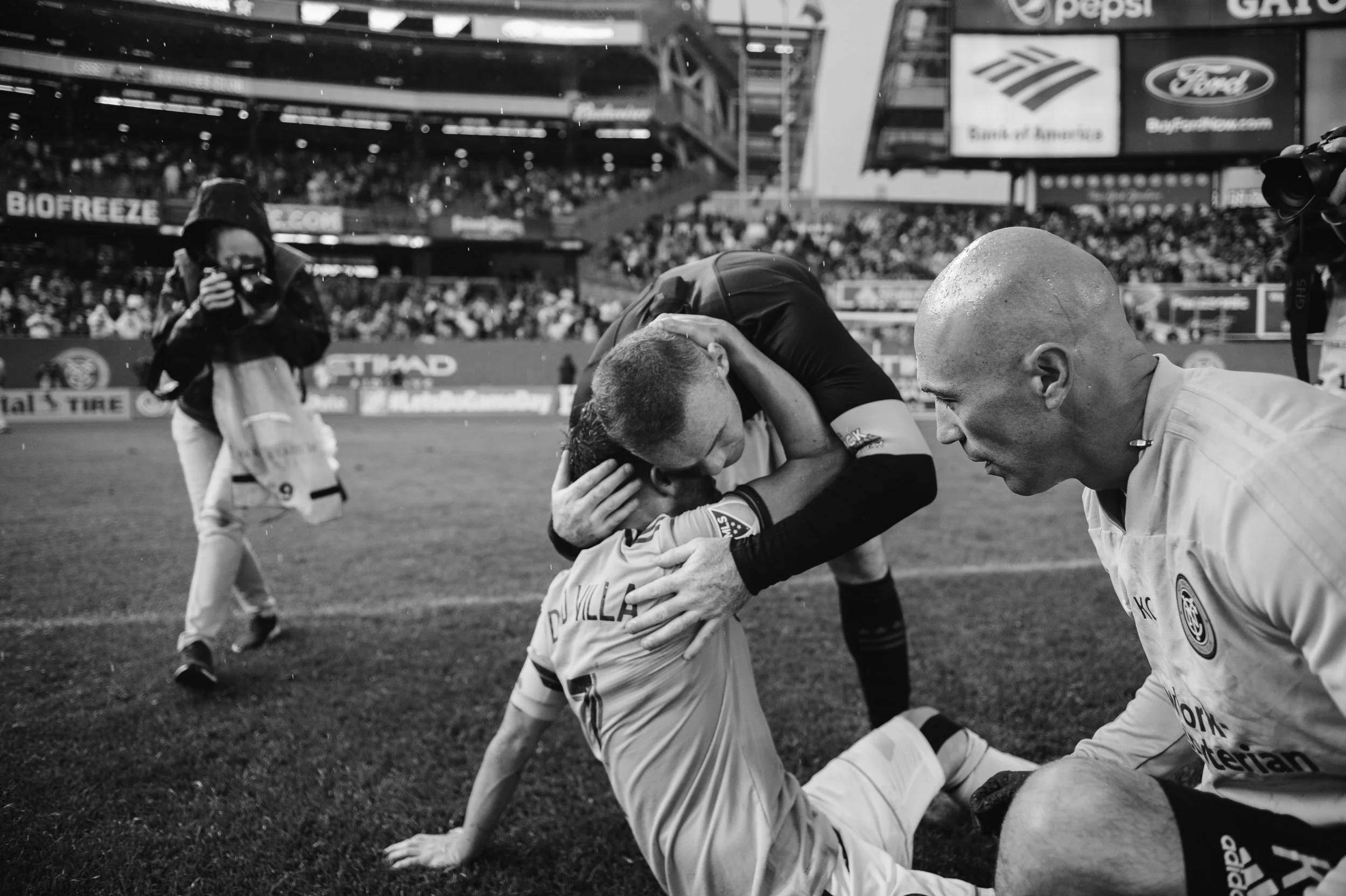 A soccer player on the ground being comforted by a teammate and a staff member after a game, with a photographer and a crowd in the background.