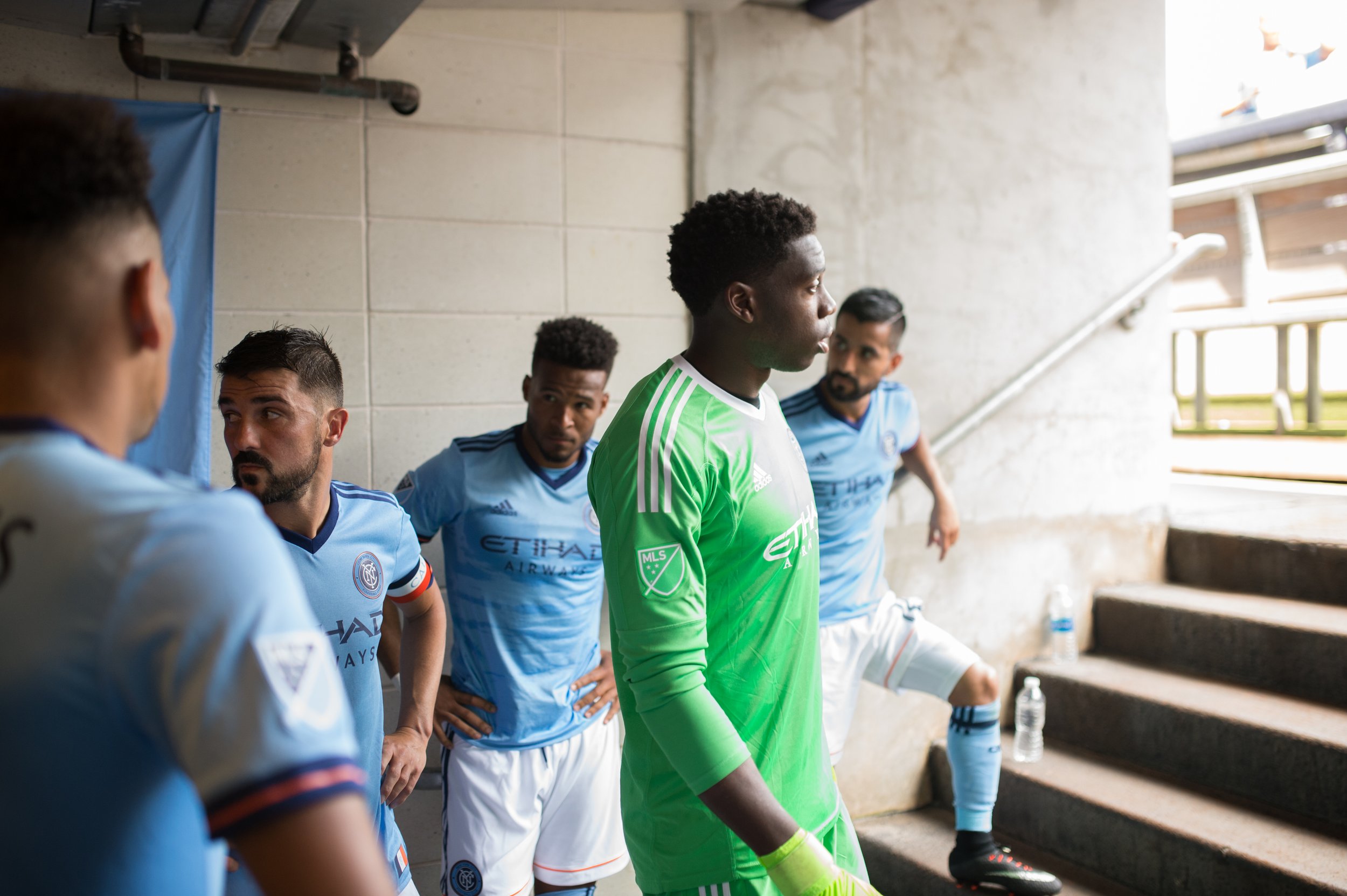 Soccer players in a tunnel before a match, with one goalie in a bright green jersey and the others in blue jerseys, some with hands on hips and listening.