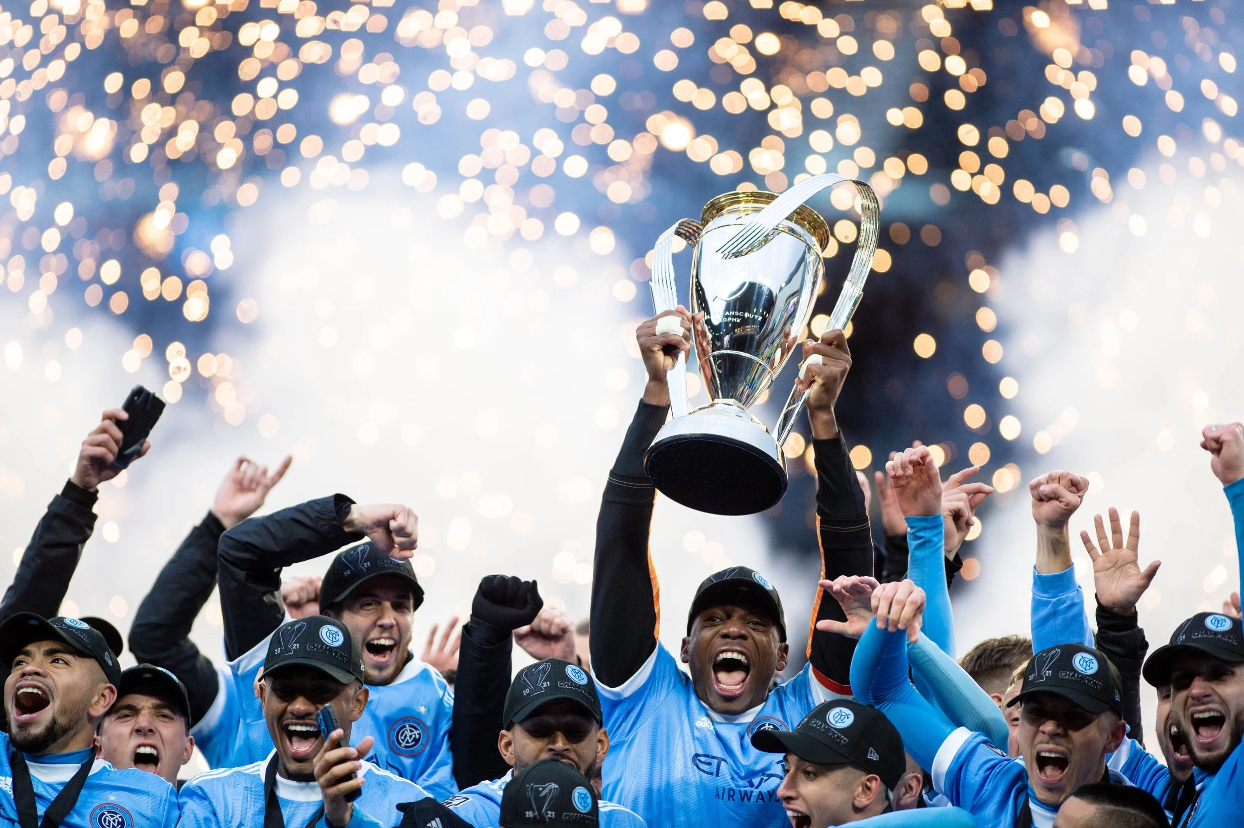 A soccer team wearing blue jerseys celebrating a victory, with one player holding a large trophy aloft, surrounded by cheering teammates, against a background of fireworks or sparkling lights.