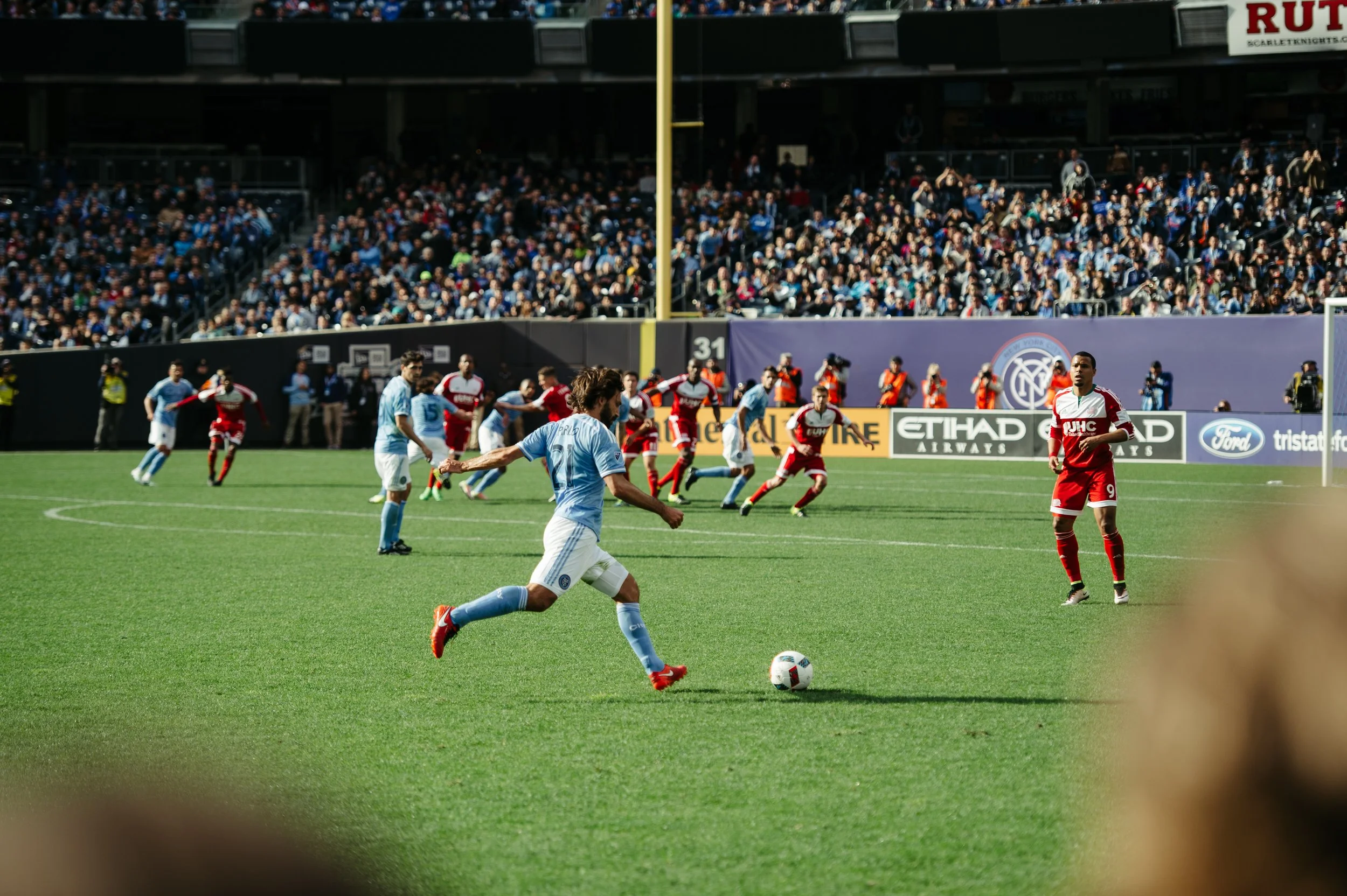 A soccer match in progress with players on the field, audience in the stands, and advertising banners, including Etihad Airways and Ford.