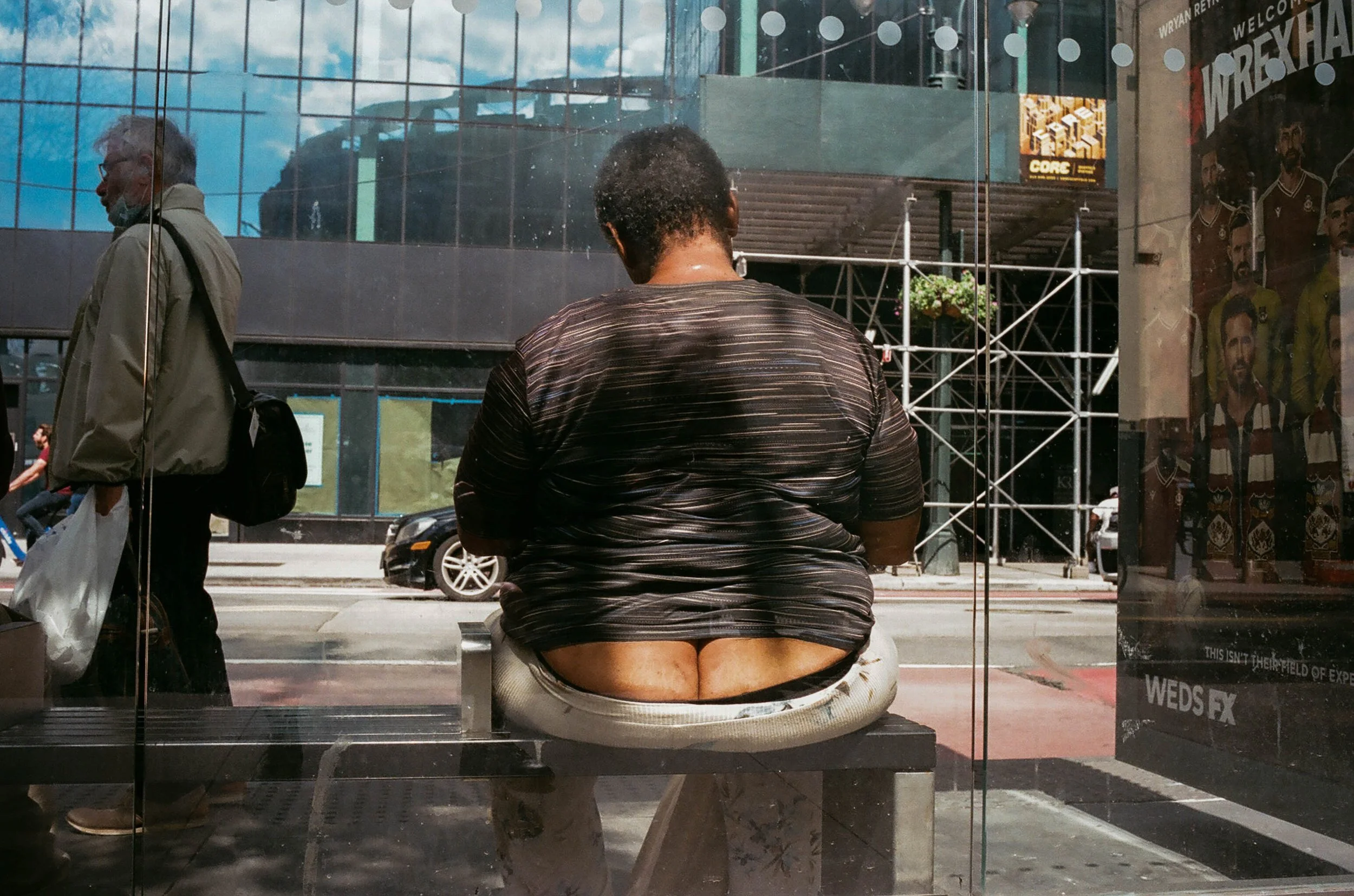 A person with short hair wearing a dark, horizontal striped shirt sitting at a bus stop, with their back to the camera. There is a reflection of a man walking past outside, and a cityscape with modern buildings and cars in the background.