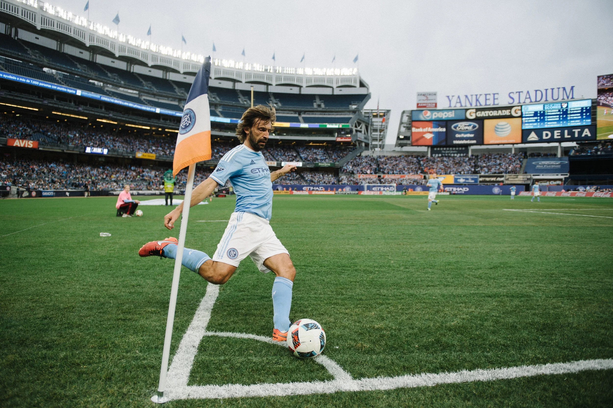 A soccer player is about to kick the ball near the corner of the field at Yankee Stadium, with the corner flag in the foreground and fans in the stands in the background.
