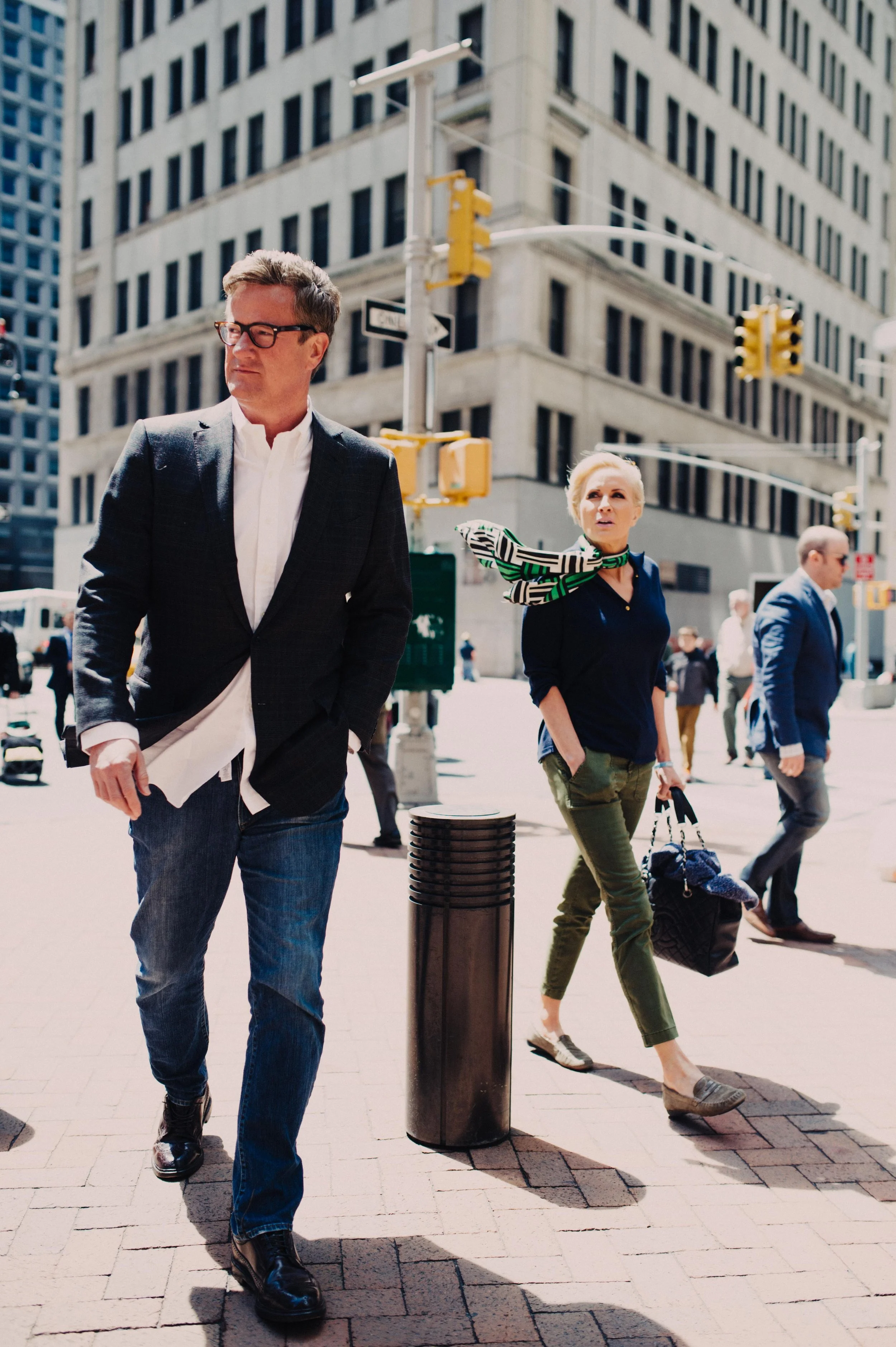 People walking on a city street corner with tall buildings, traffic lights, and a cityscape in the background.