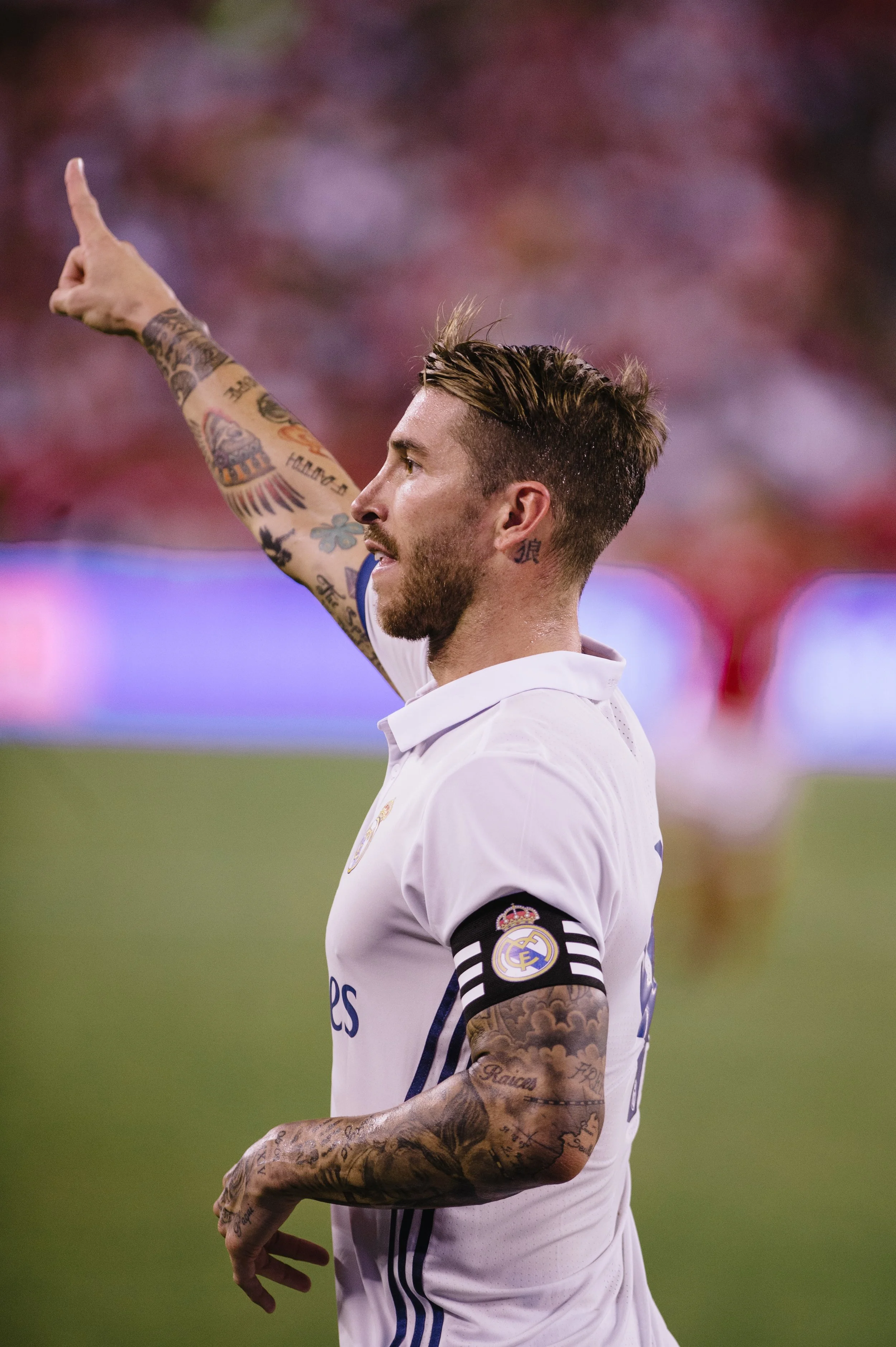 A football player in a white jersey with the Real Madrid logo, raising his right arm with his index finger pointing upward during a game.