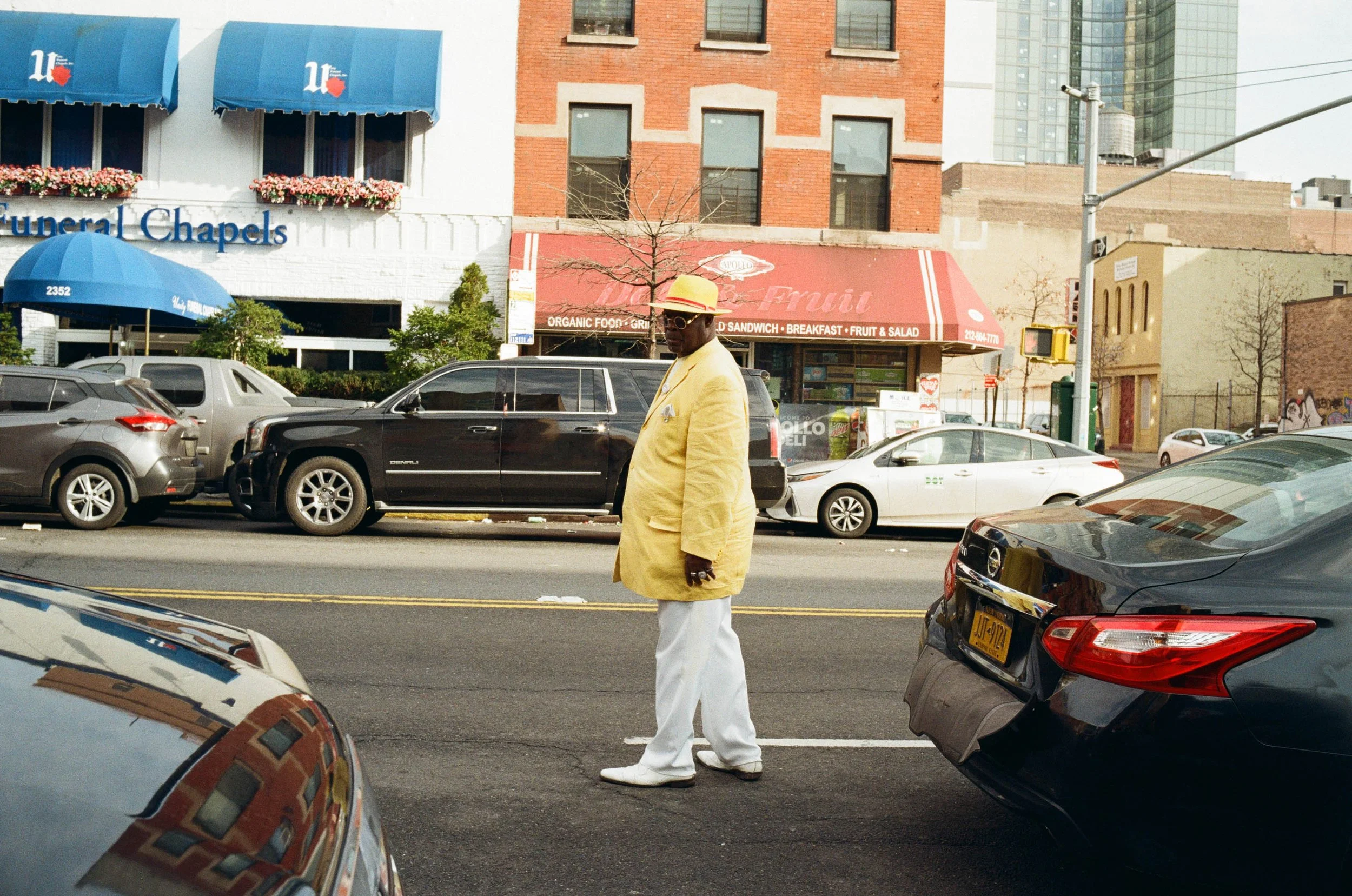 A man dressed in a bright yellow coat, yellow hat, white pants, and white shoes walking across a city street. Cars are parked along the street, and buildings with storefronts, including a funeral chapel and a fruit shop, line the background.