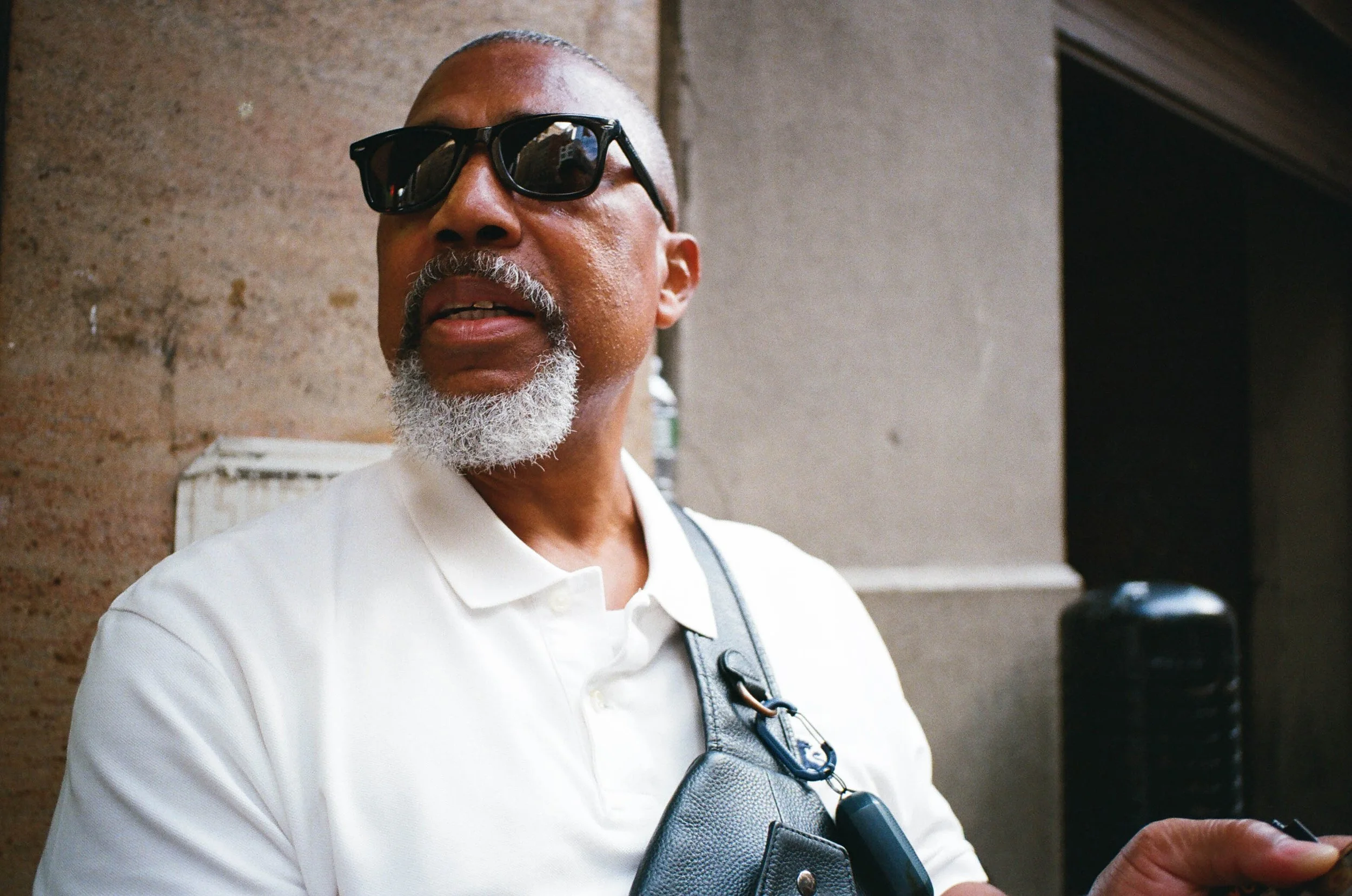 An older man wearing dark sunglasses and a white collared shirt, with a gray beard, standing in front of a stone wall.
