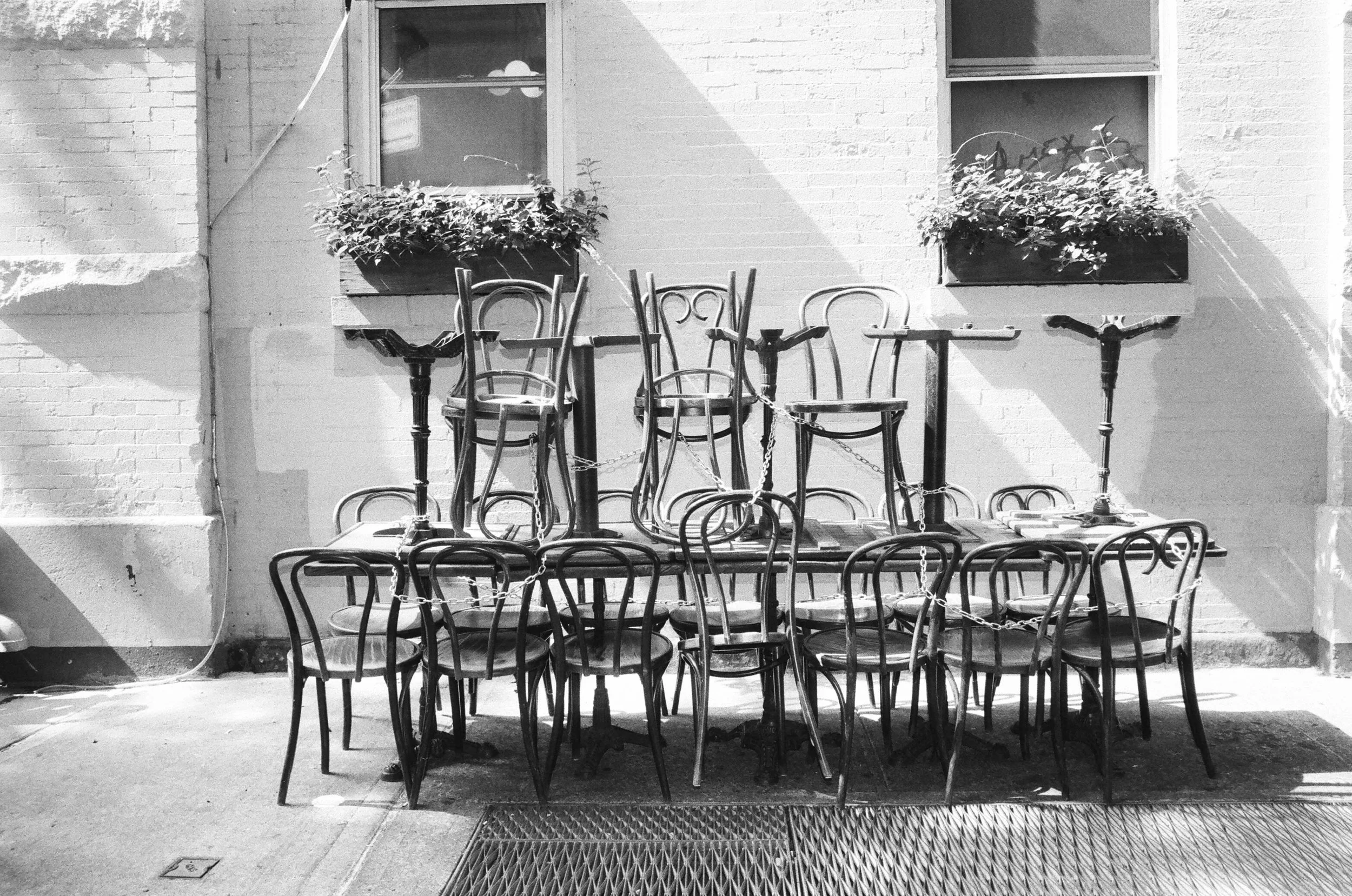 Stacked chairs and tables outdoors, chained together, against a brick wall with two windows and flower boxes.