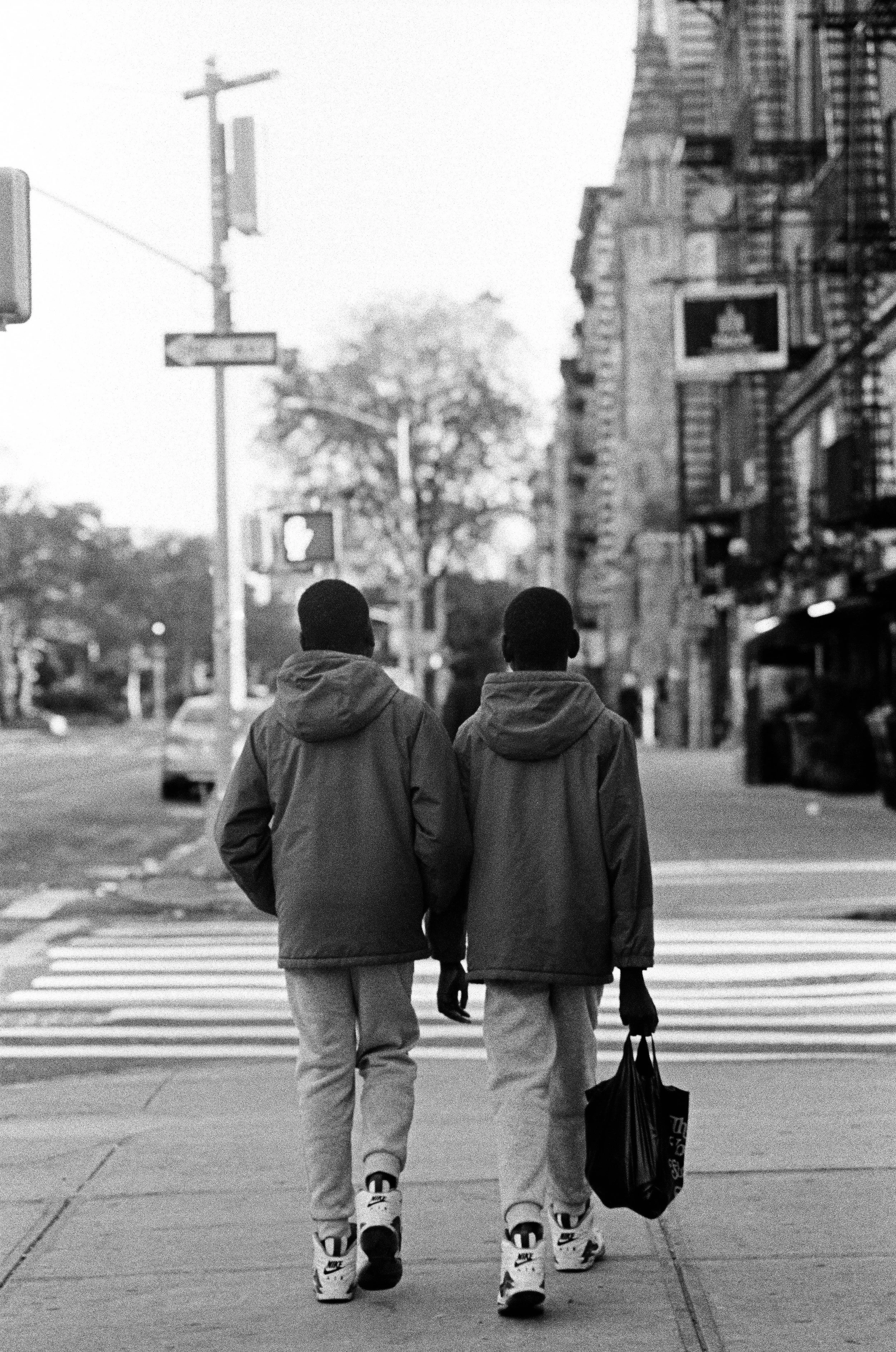 Two young men walking away from the camera on a city sidewalk, dressed in hoodies and jackets, with a shopping bag in one hand, black and white photo.