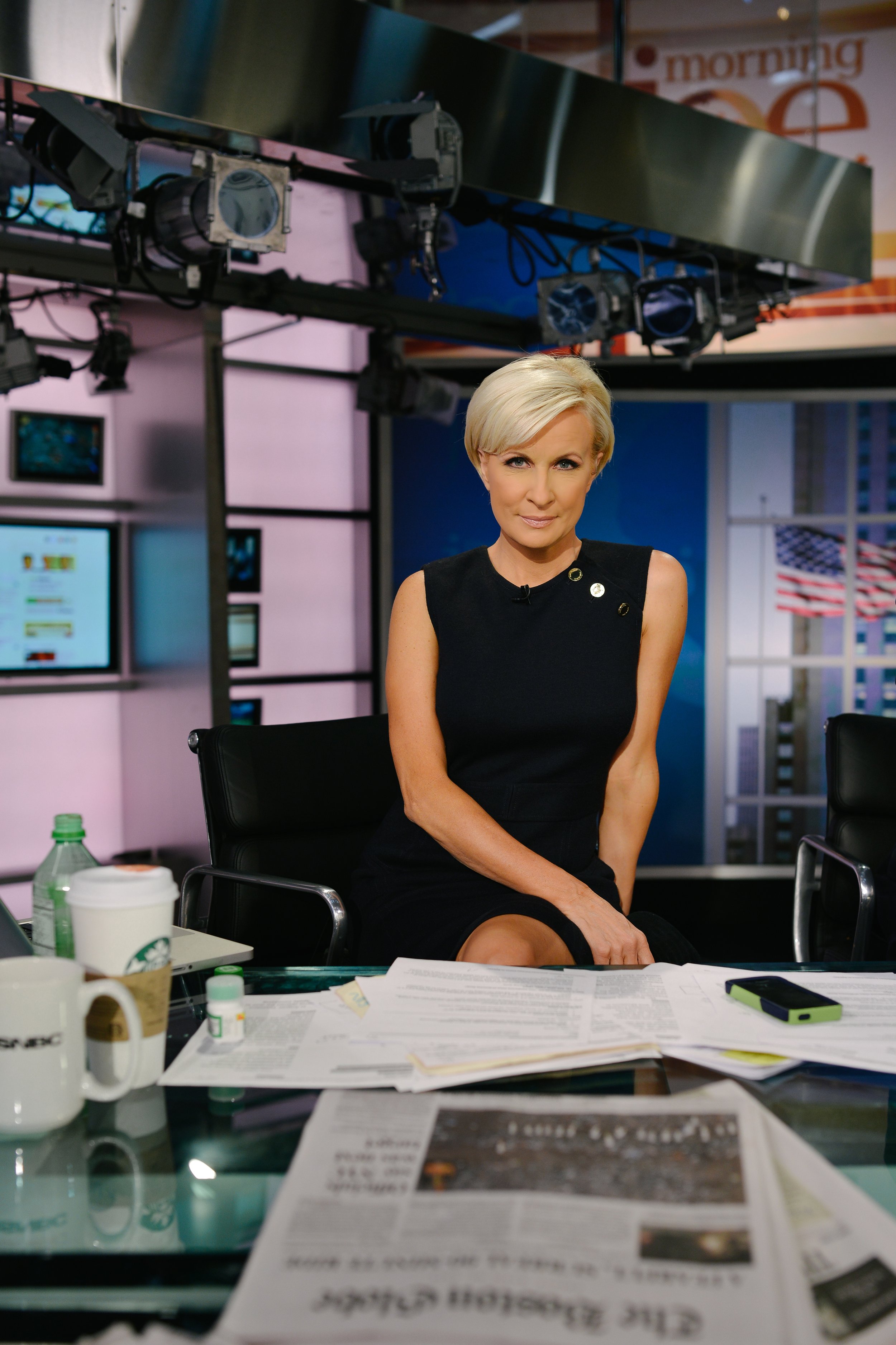 A female news anchor with short blonde hair sitting at a news desk in a studio, with papers, coffee cups, and a phone on the desk, and multiple monitors and studio equipment in the background.