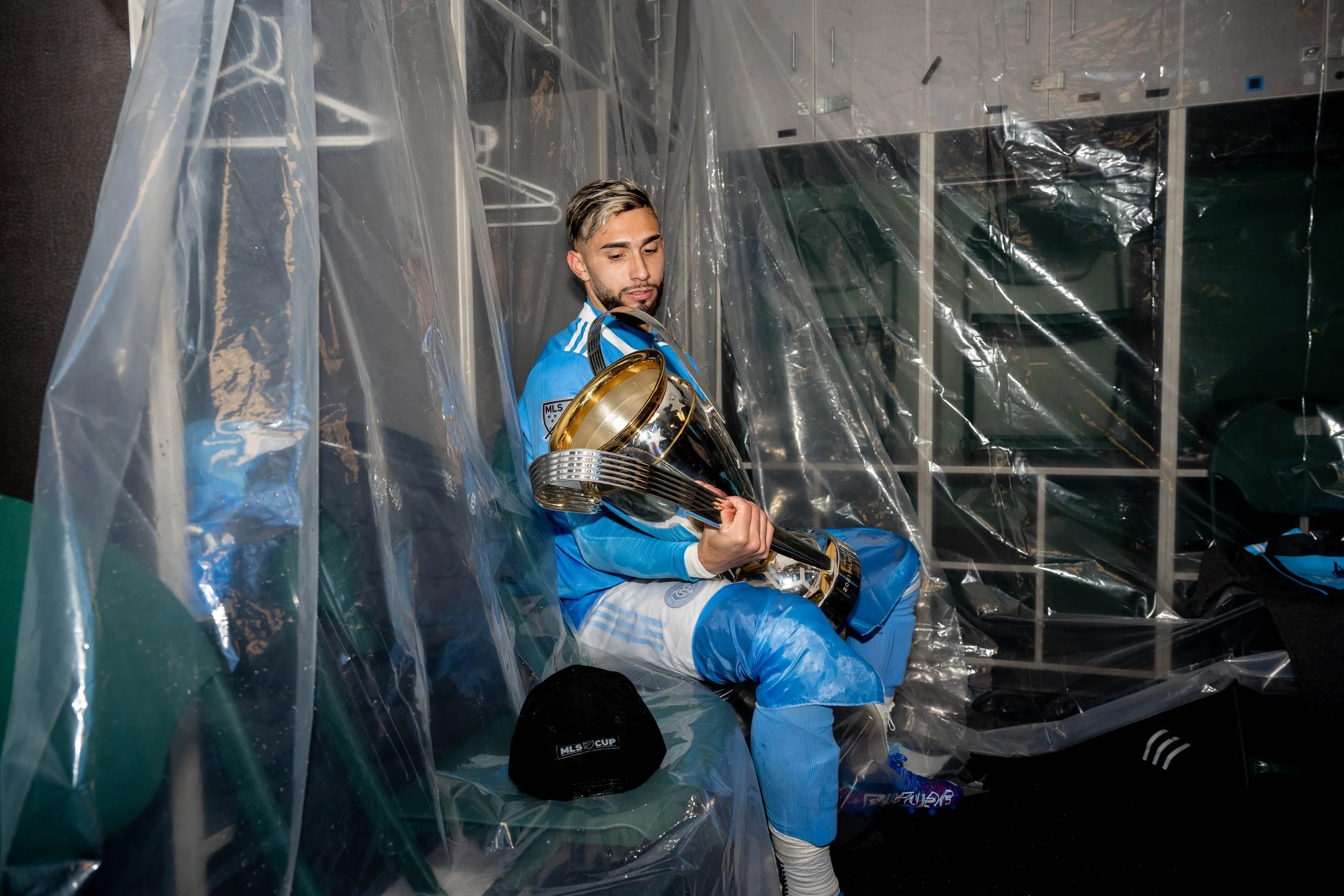 A male soccer player in a blue uniform sitting on a bench inside a locker room, holding a large trophy. The room is covered with clear plastic and has lockers and a black cap with 'MLS Cup' written on it placed on the bench.