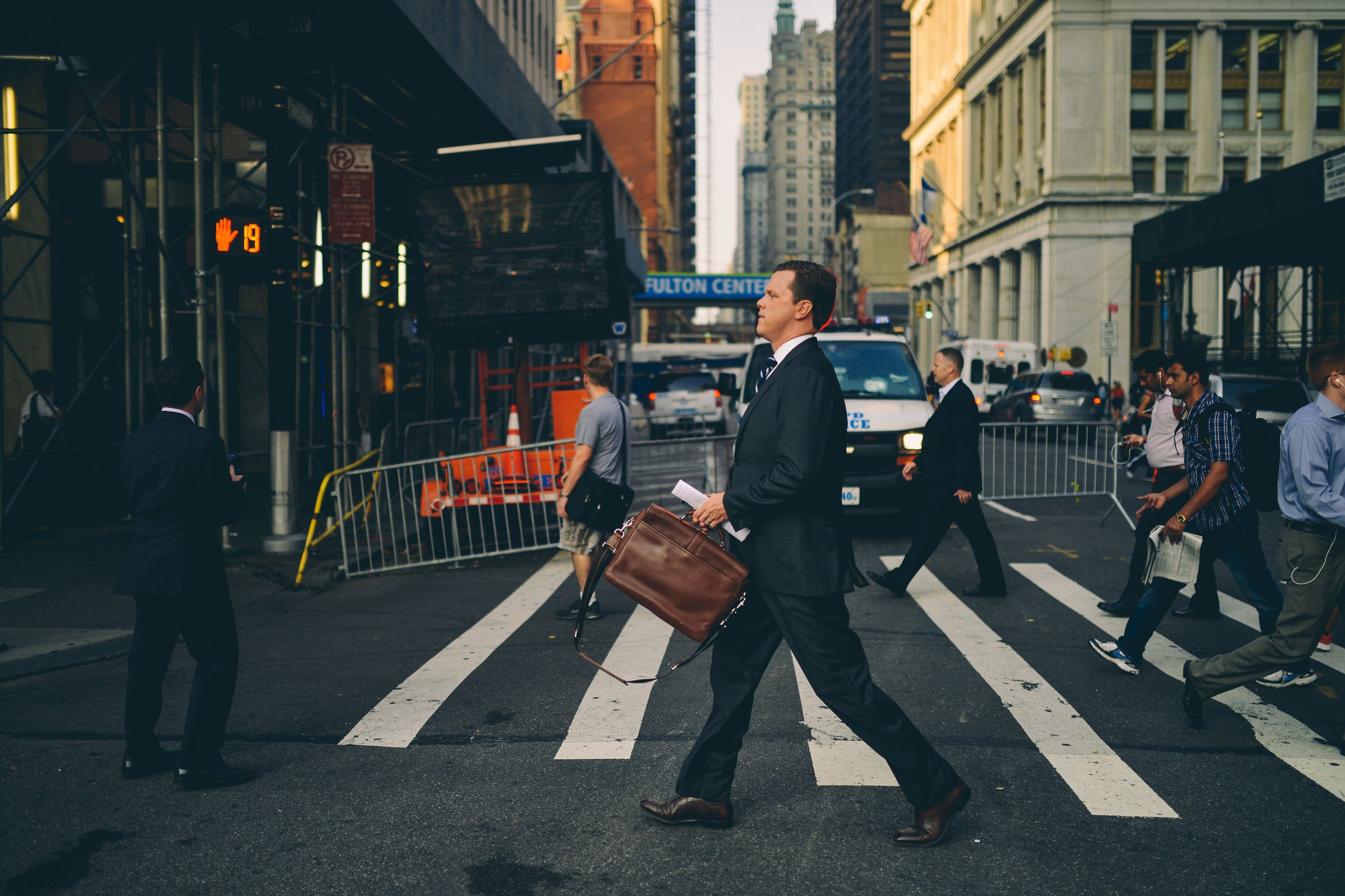 Business professionals and pedestrians crossing a city street in front of buildings and vehicles, with a prominent man in the center carrying a briefcase and walking.