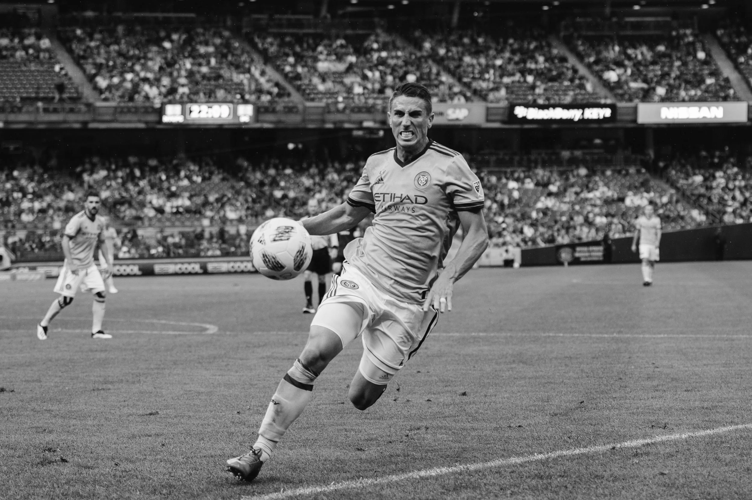 A soccer player in a New York City FC jersey yelling and running on the field with a ball, with two teammates and a crowd in the background.