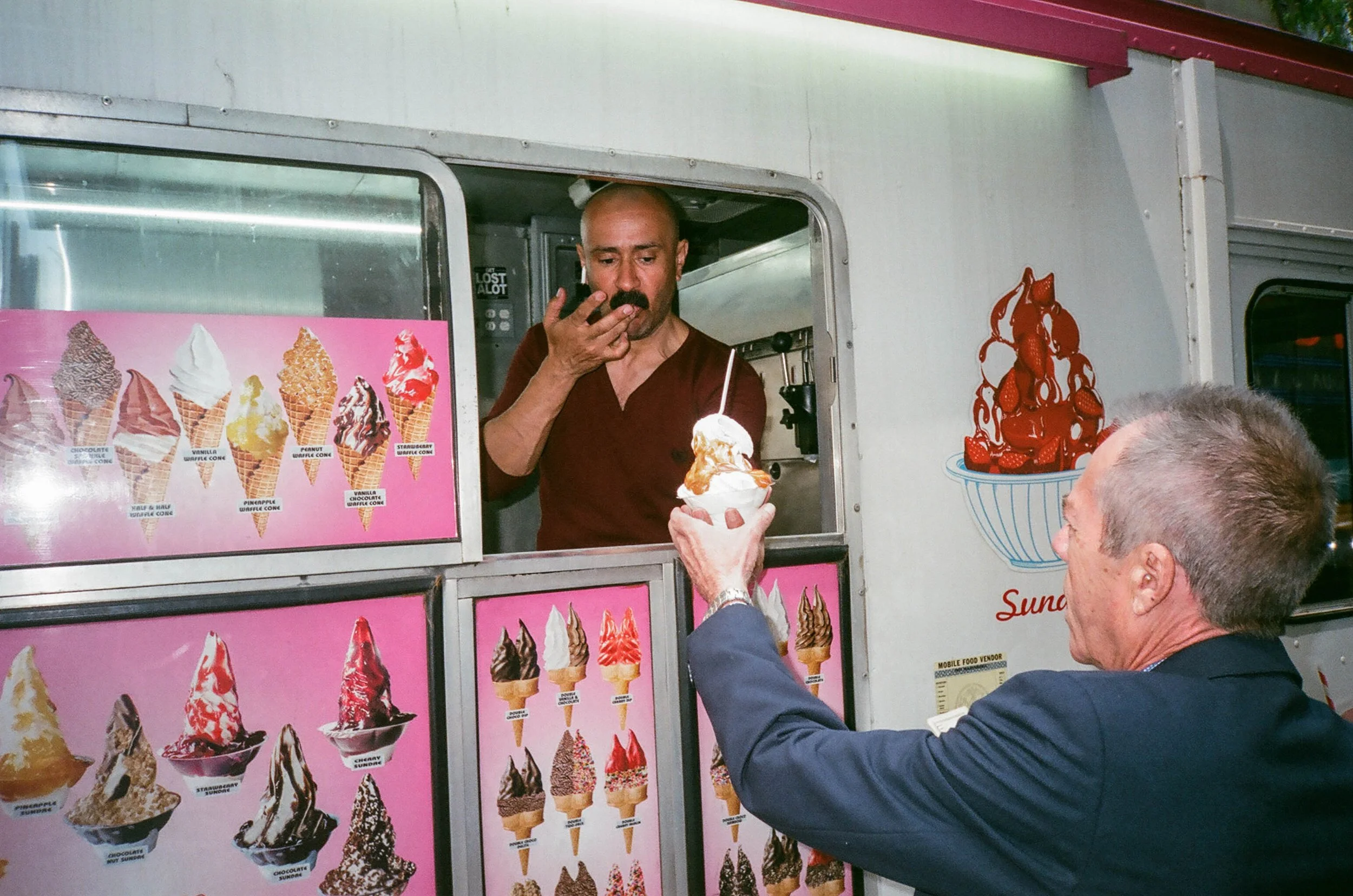 A man in a dark suit is handing a cup of ice cream to a man working at an ice cream truck, which displays various flavors of soft serve ice cream in pictures on pink backgrounds.