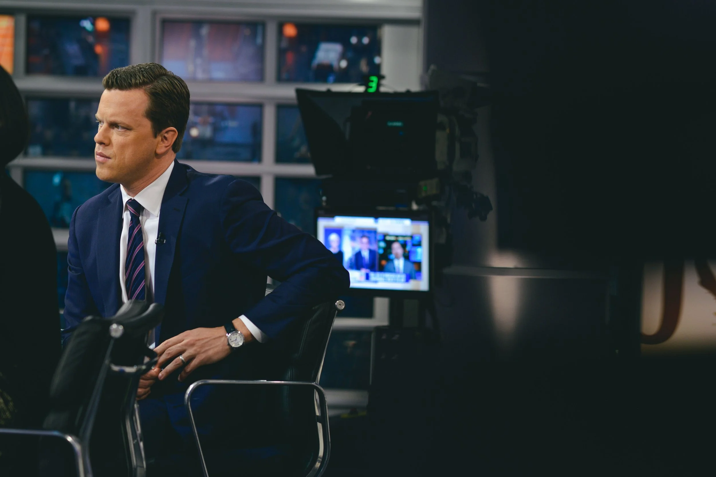 A man in a blue suit and striped tie sitting in a news studio, appearing serious, with television screens and cameras in the background.