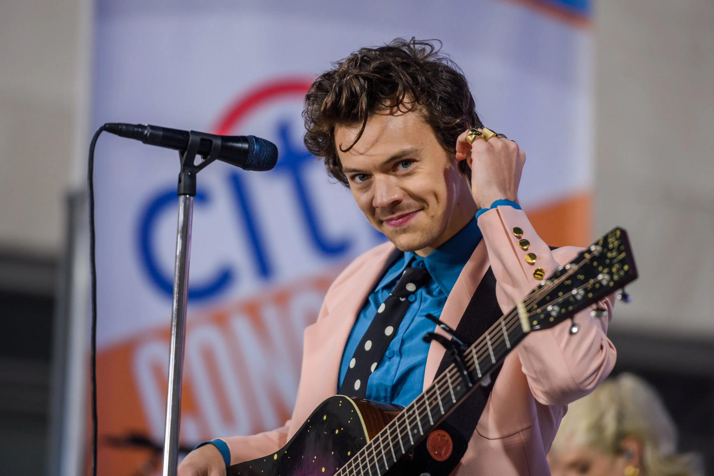 A young man with curly hair, wearing a pink suit jacket with star-shaped buttons, a blue shirt, and a polka dot tie, holding a guitar and smiling at the camera.