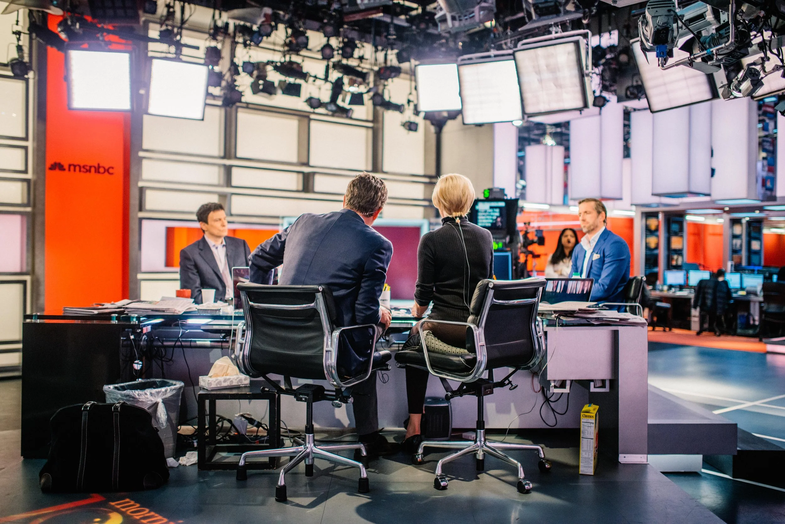 Television news studio with five professionals around a table, multiple screens, lighting, and camera equipment.