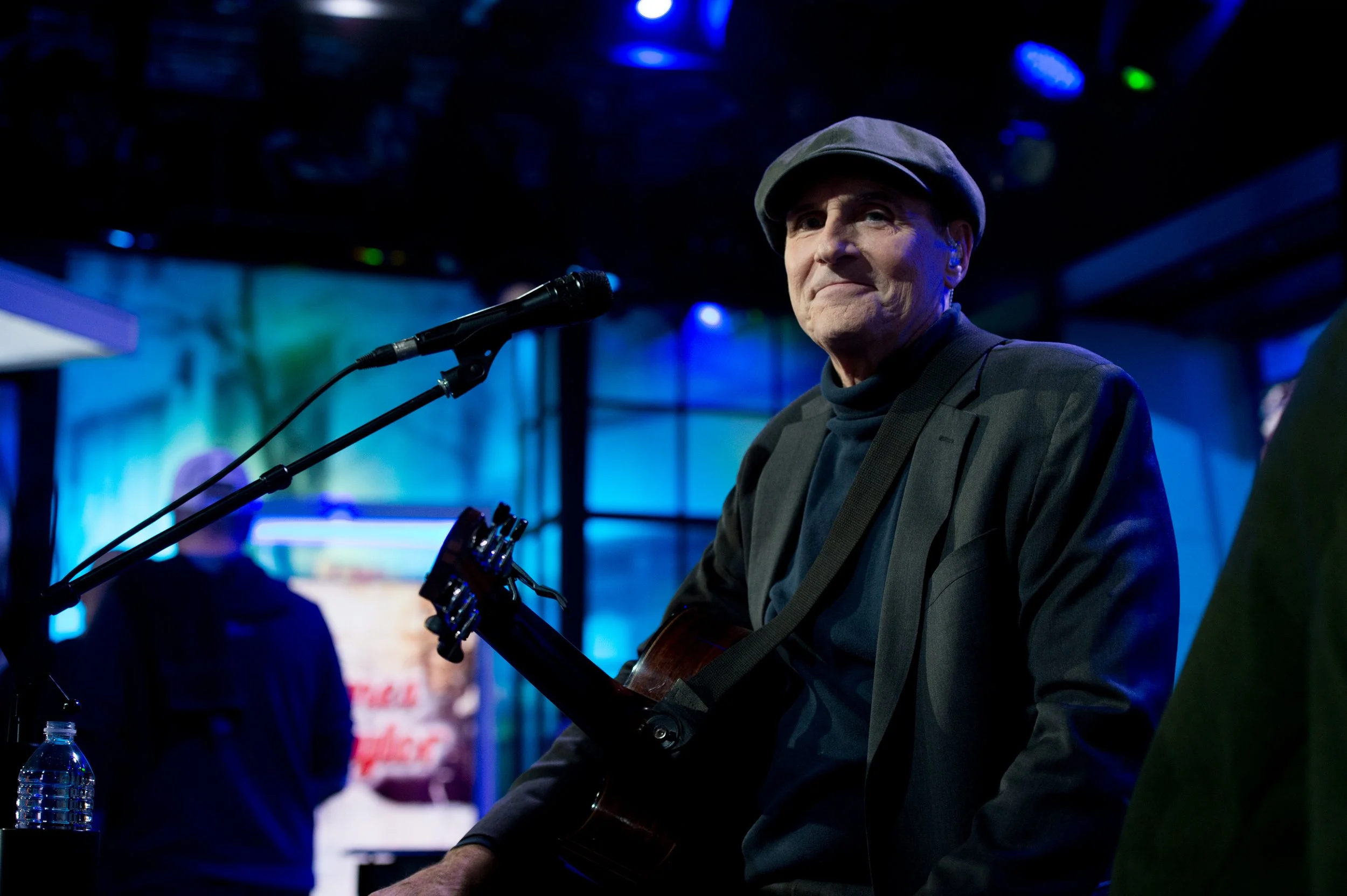 A man wearing a flat cap and dark clothing playing an acoustic guitar on stage, with a microphone in front of him, in a dimly lit venue with blue and green stage lights.