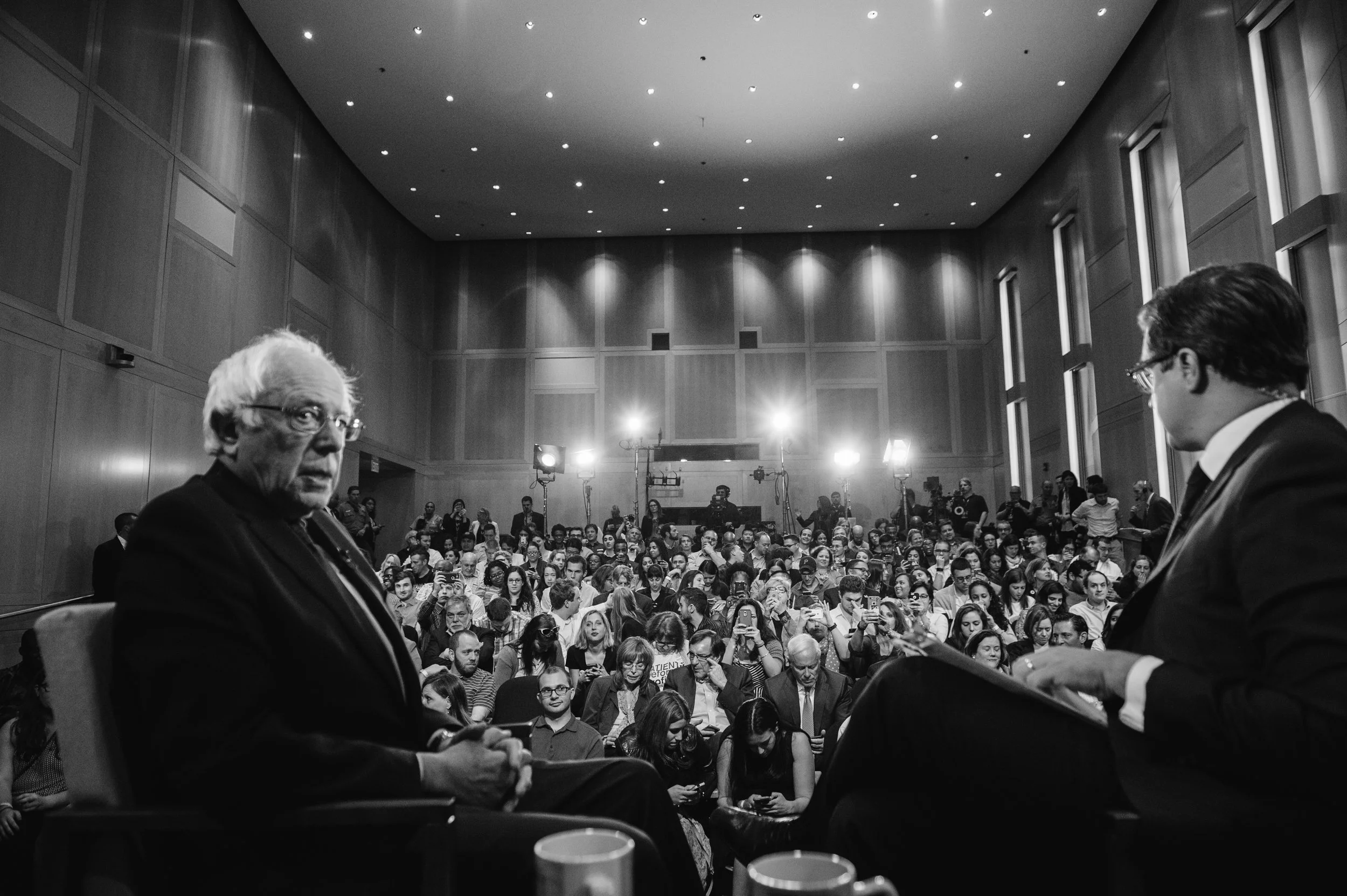 Two men in suits seated on stage during a panel discussion with a large audience in a conference hall.