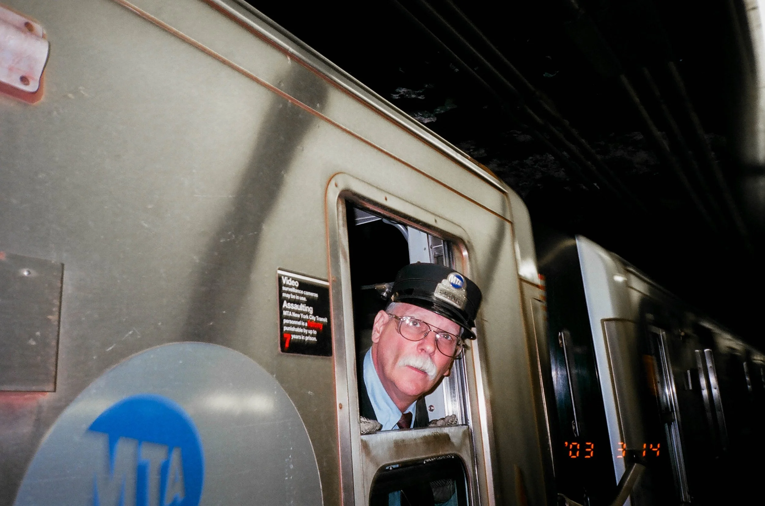 An older man with a white mustache and glasses, wearing a black conductor's hat, leans out of a subway train window.