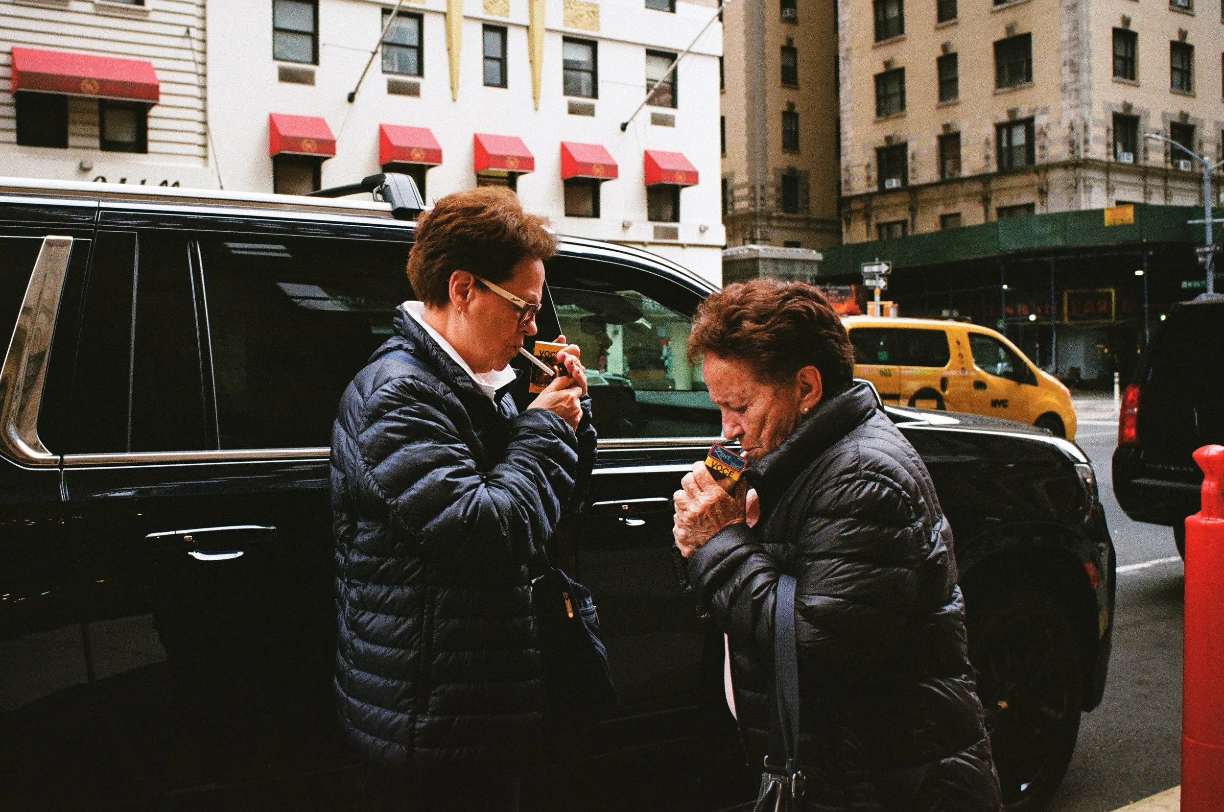 Two women standing on a city street in front of a black SUV, engaging in an activity with small electronic devices or cards, with yellow taxis and tall buildings in the background.