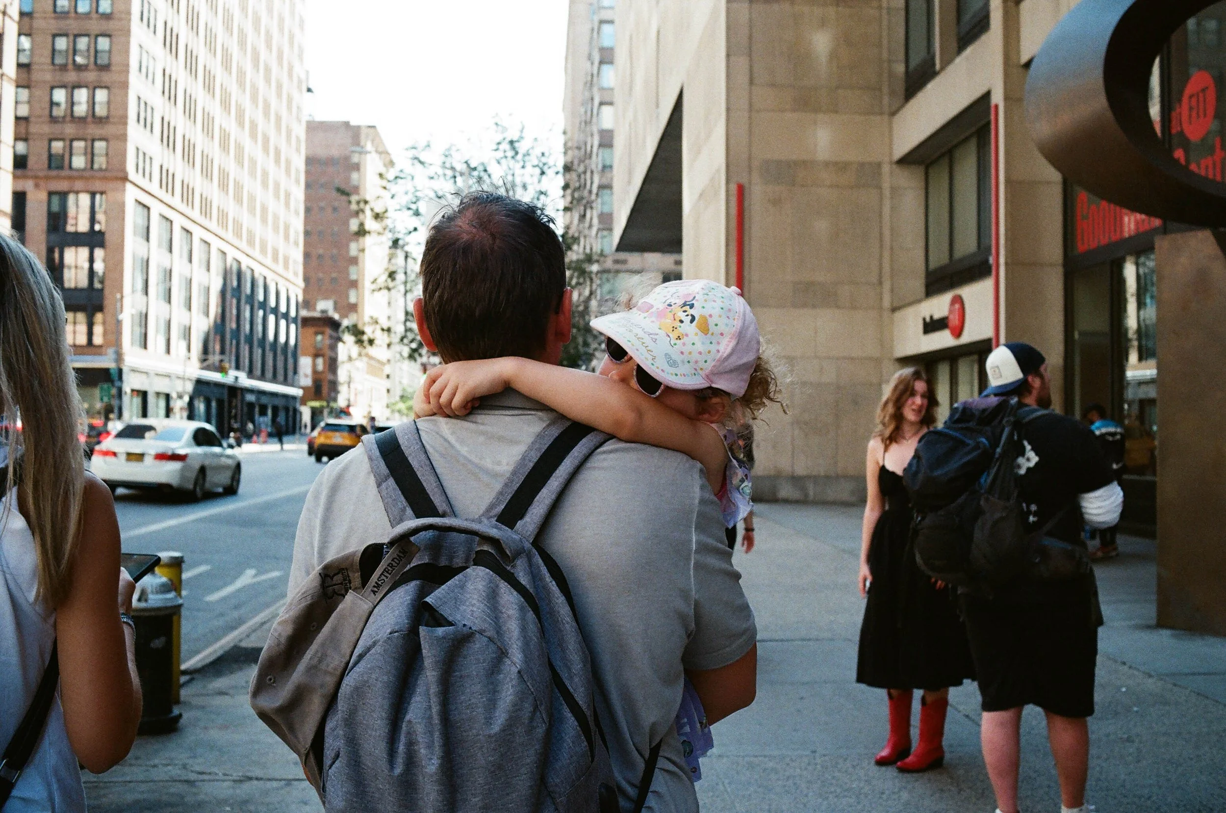 A man carrying a young girl with curly hair, wearing sunglasses and a colorful hat, on a city sidewalk. The girl has her arms around his neck. People are walking by, including a woman in a black dress and a man with a backpack. Cars and tall building