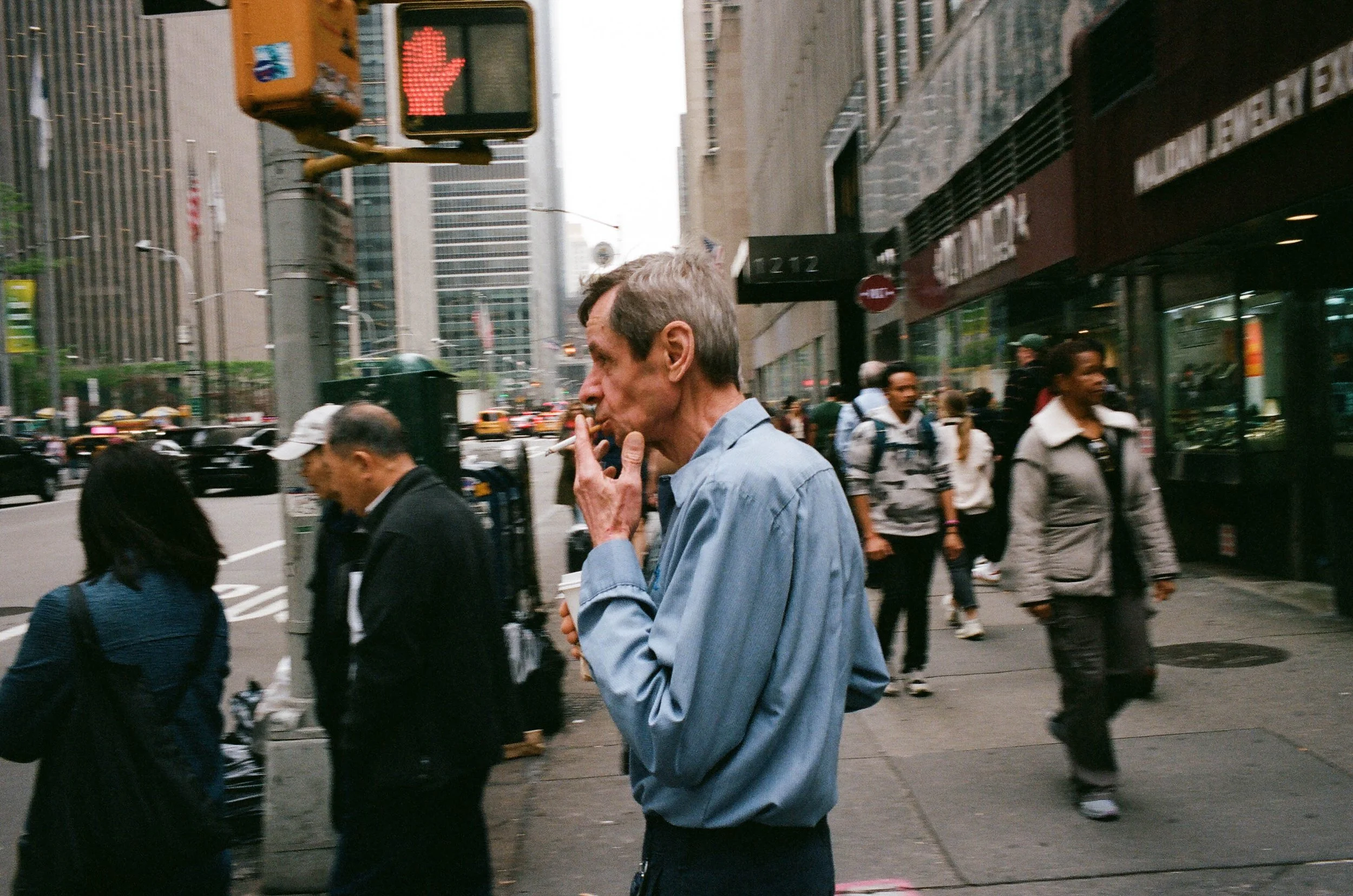 A man with gray hair and a blue shirt stands on a bustling city sidewalk, smoking a cigarette and holding a coffee cup, with tall buildings and pedestrians in the background.