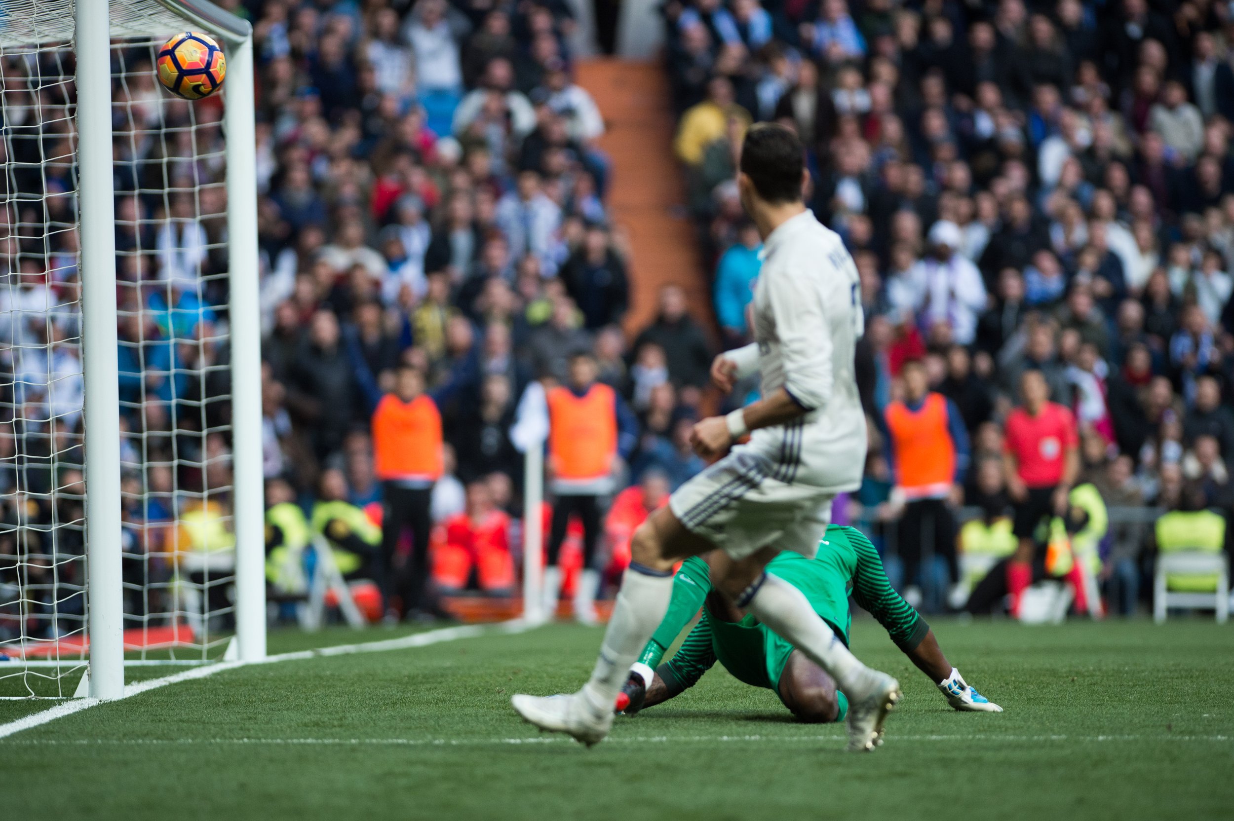 A soccer player in a white uniform kicking a ball towards the goal, with a goalkeeper in green on the ground and a crowd watching in the background.