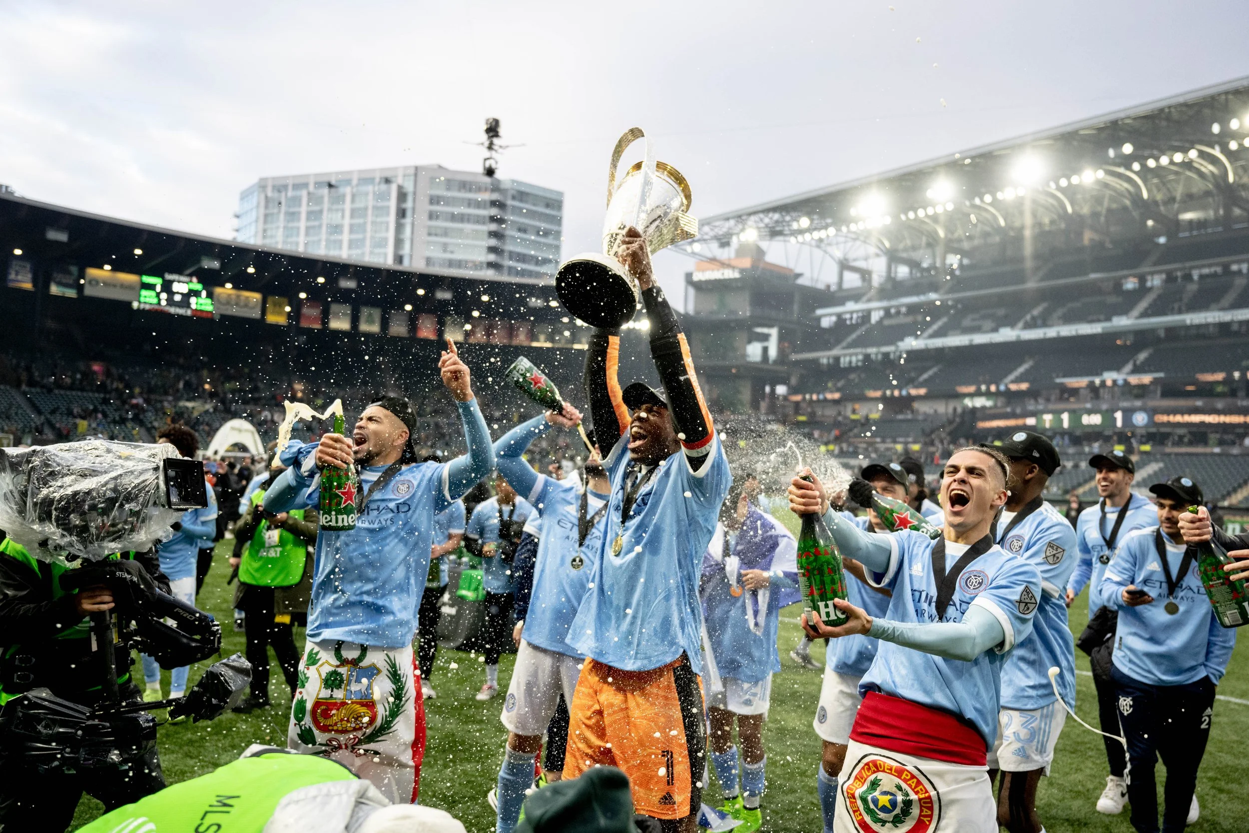 Soccer players celebrating on the field after winning a match, spraying champagne and wearing medals, with a stadium in the background.